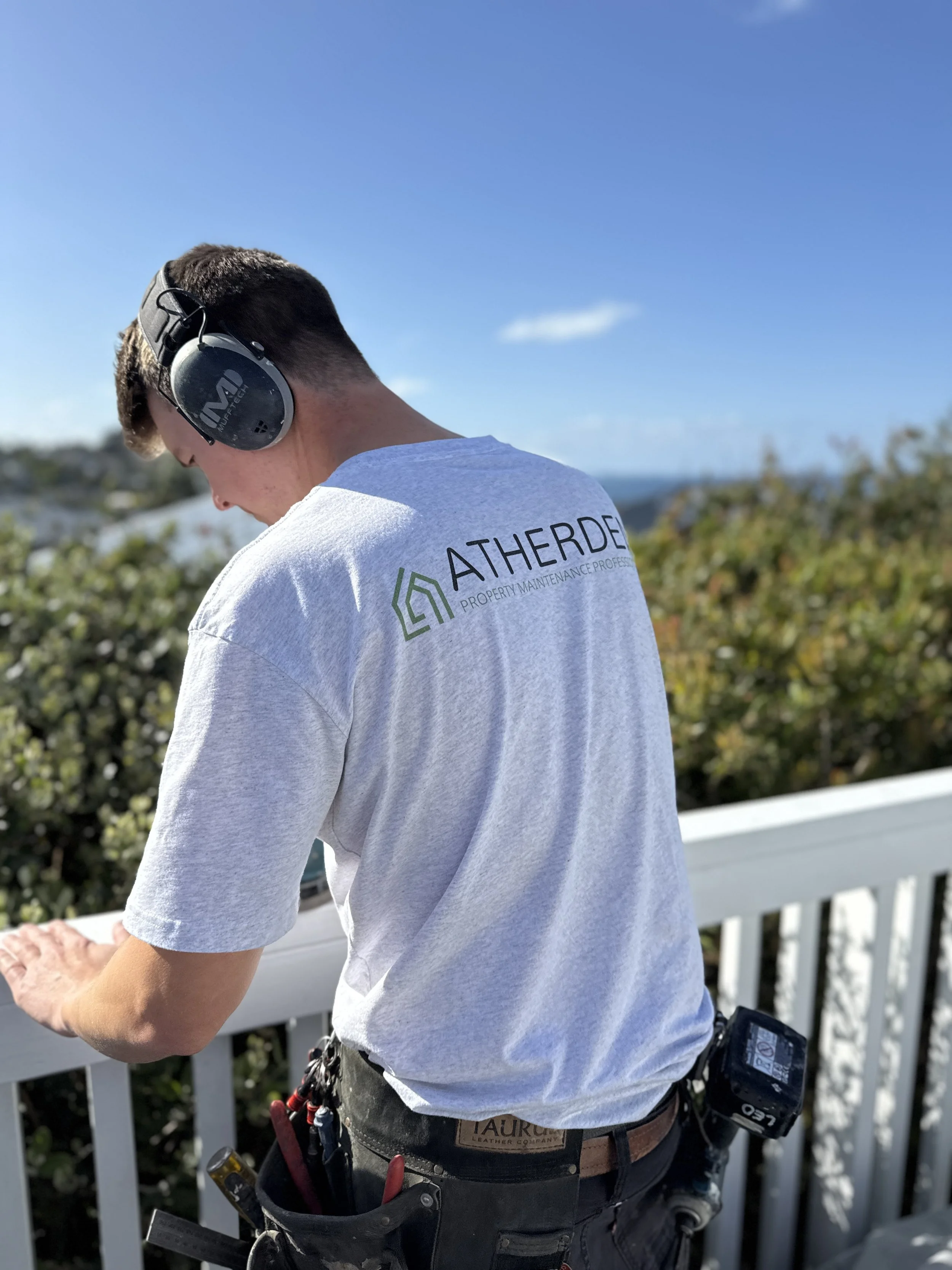 A man in a gray T-shirt with the text ''ATHERDE PROPERTY MAINTENANCE PROFESSIONAL'' on the back, wearing headphones, working outdoors on a white railing, with trees, bushes, and a blue sky in the background.