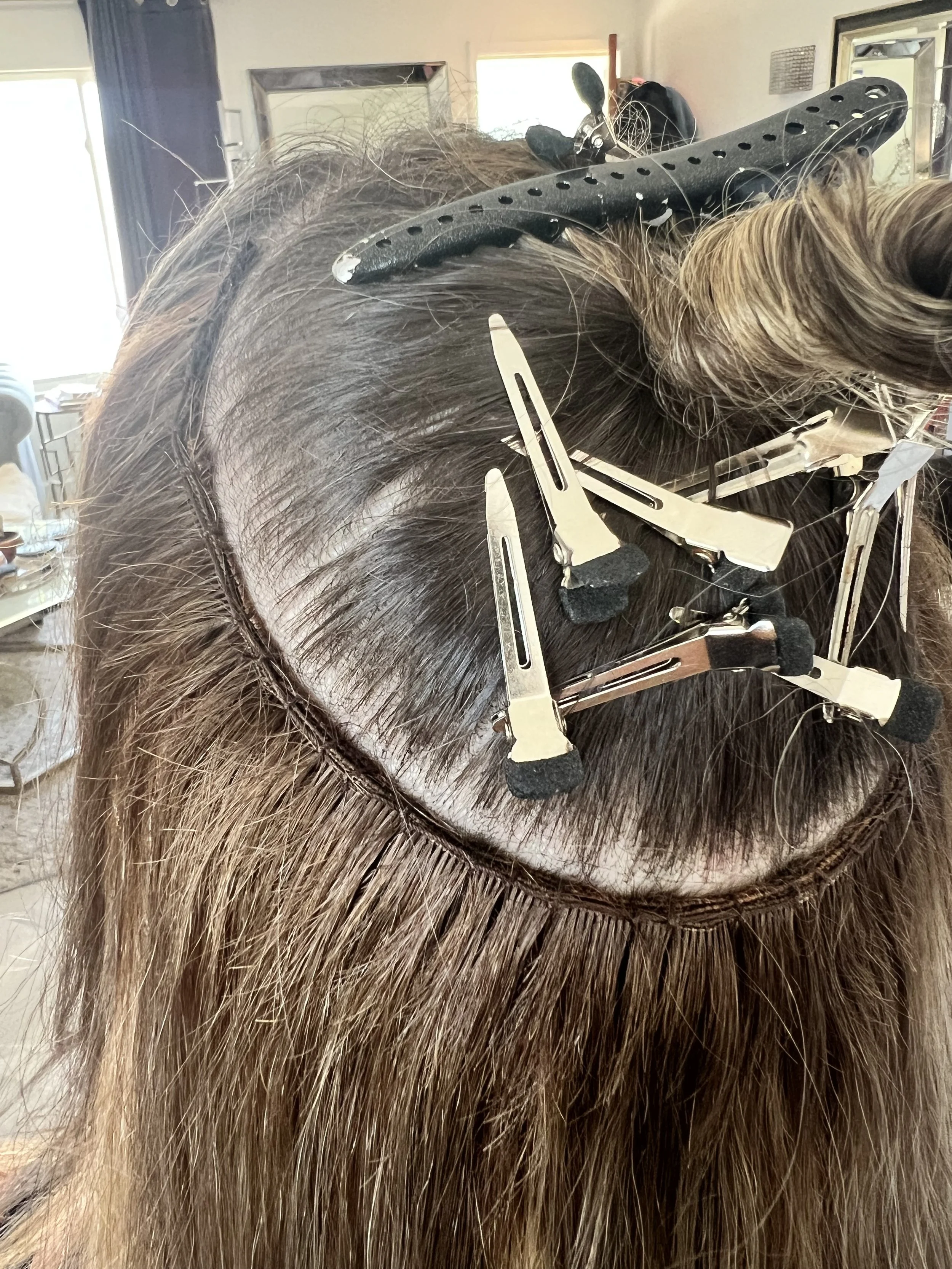 A person undergoing hair extension installation with hair clips and clips holding the hair in place, inside a room with mirrors and windows.