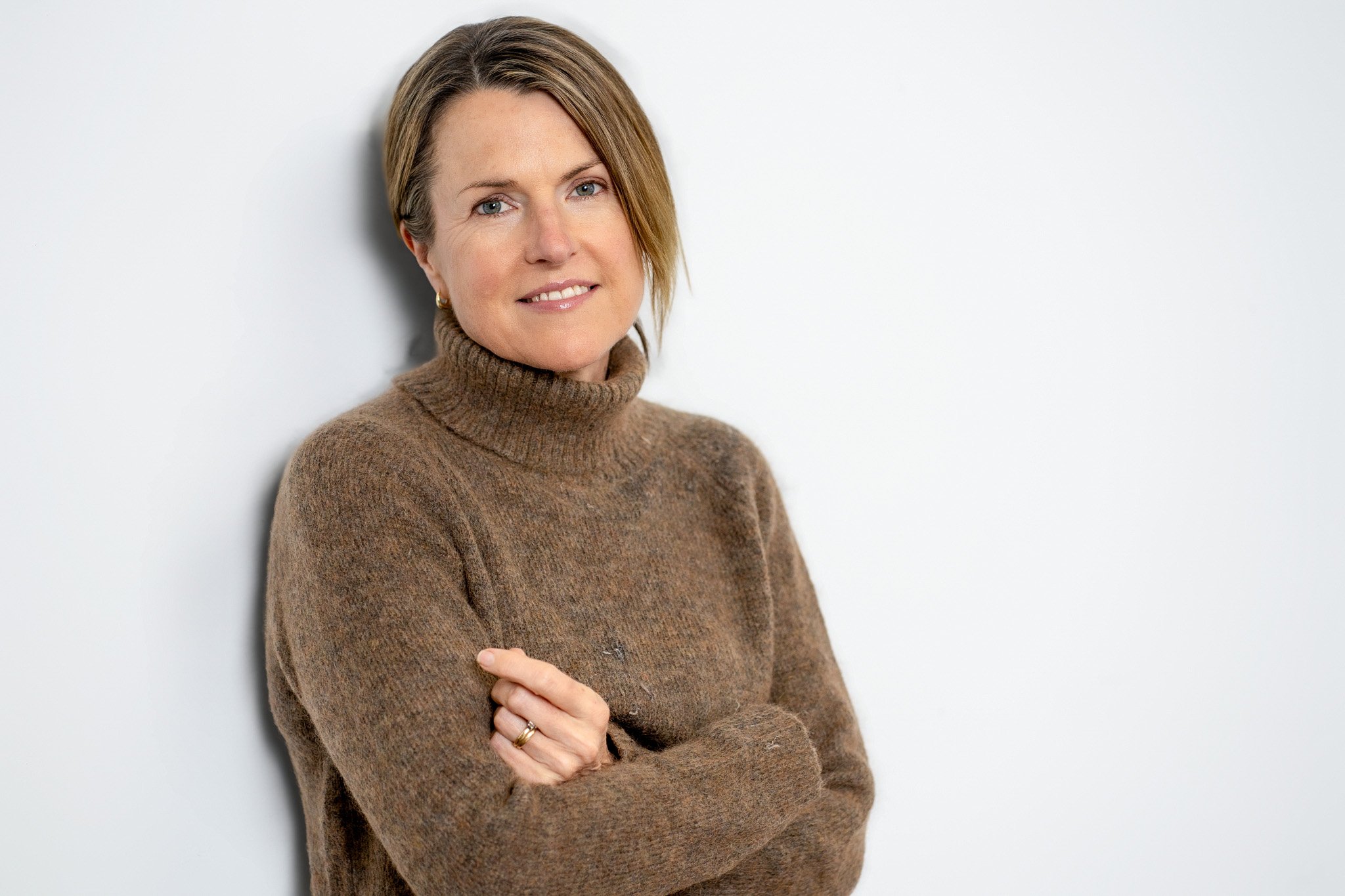 A woman with short brown hair wearing a brown turtleneck sweater, standing against a plain white background, smiling gently, with her arms crossed.