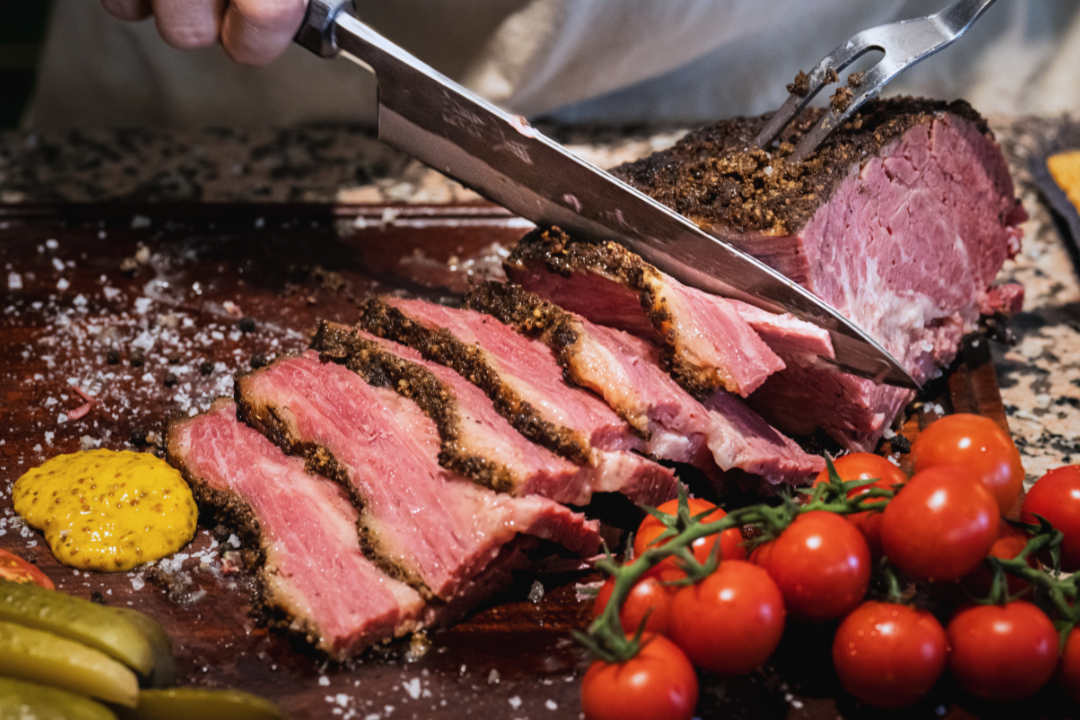 A person slicing a large hunk of zaidys brisket pastrami, seasoned with spices on a wooden cutting board, with cherry tomatoes and mustard on the side.