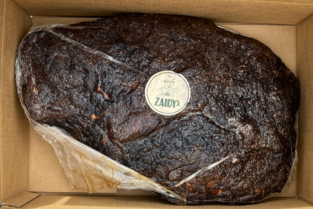 Large pastrami brisket hunk with a charred, dark crust in a cardboard box, labeled with a sticker that reads 'Zaidys' and features the zaidys grandpa character.