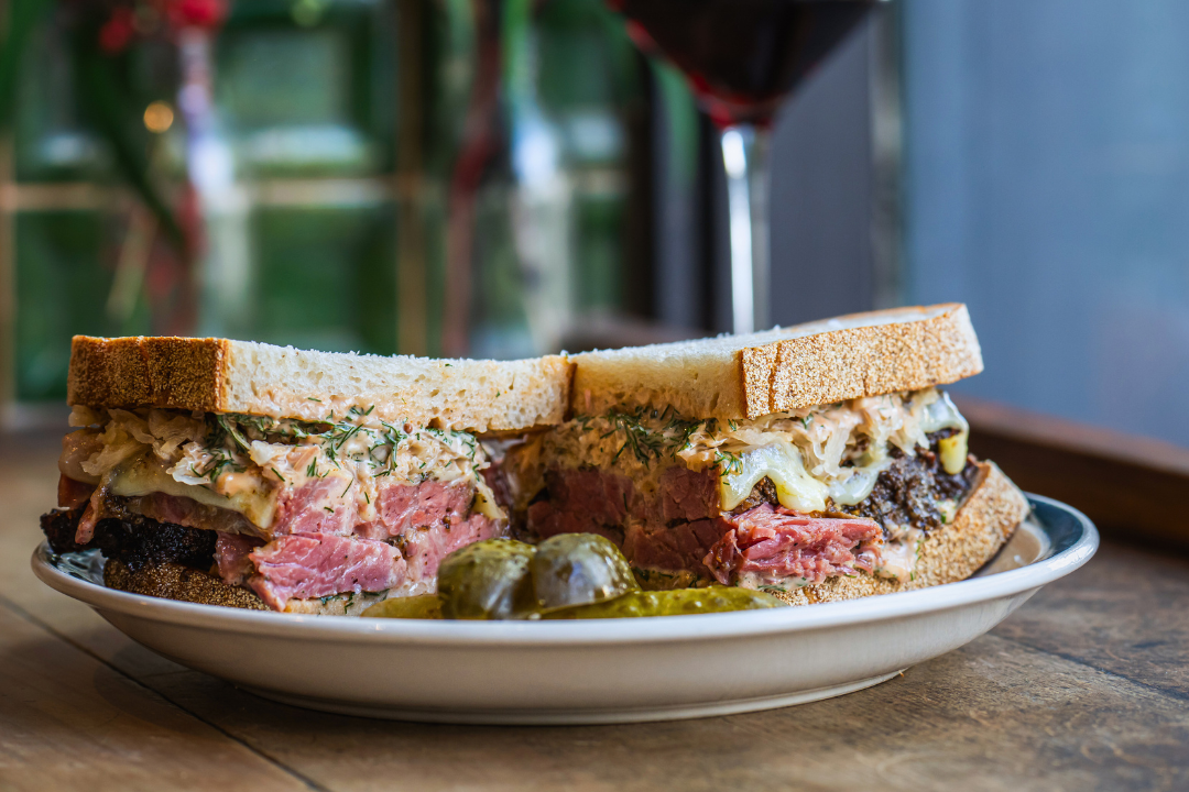 Close-up of a pastrami reuben sandwich on a white plate with pickles and a drink in the background.