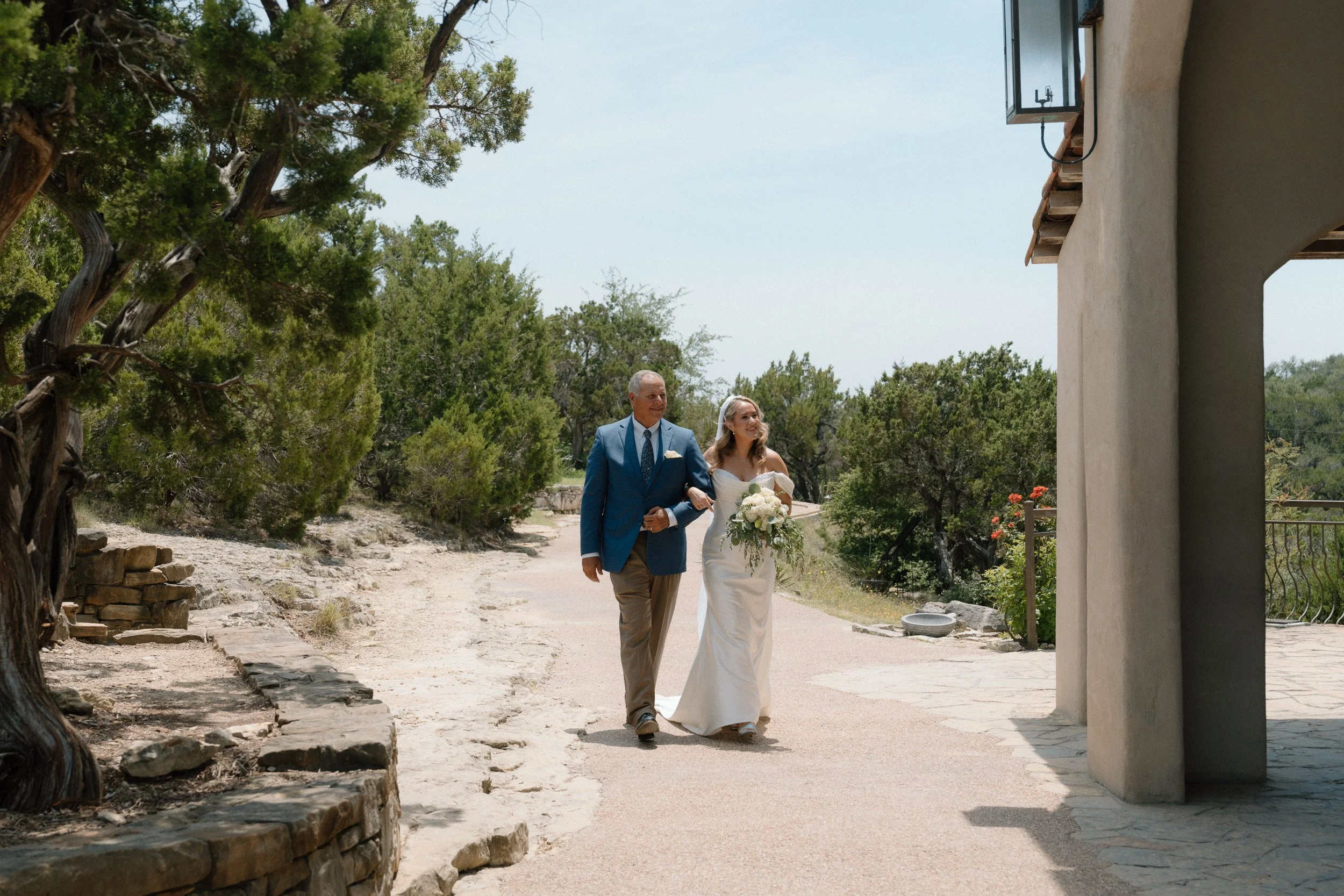 Bride and her father walk toward the chapel as she carries a white and greenery bouquet