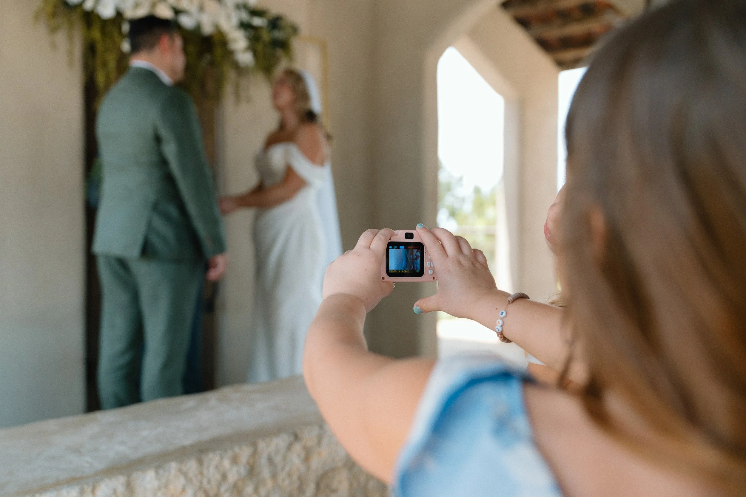 A young guest captures the ceremony on a small pink camera as the couple stands at the altar