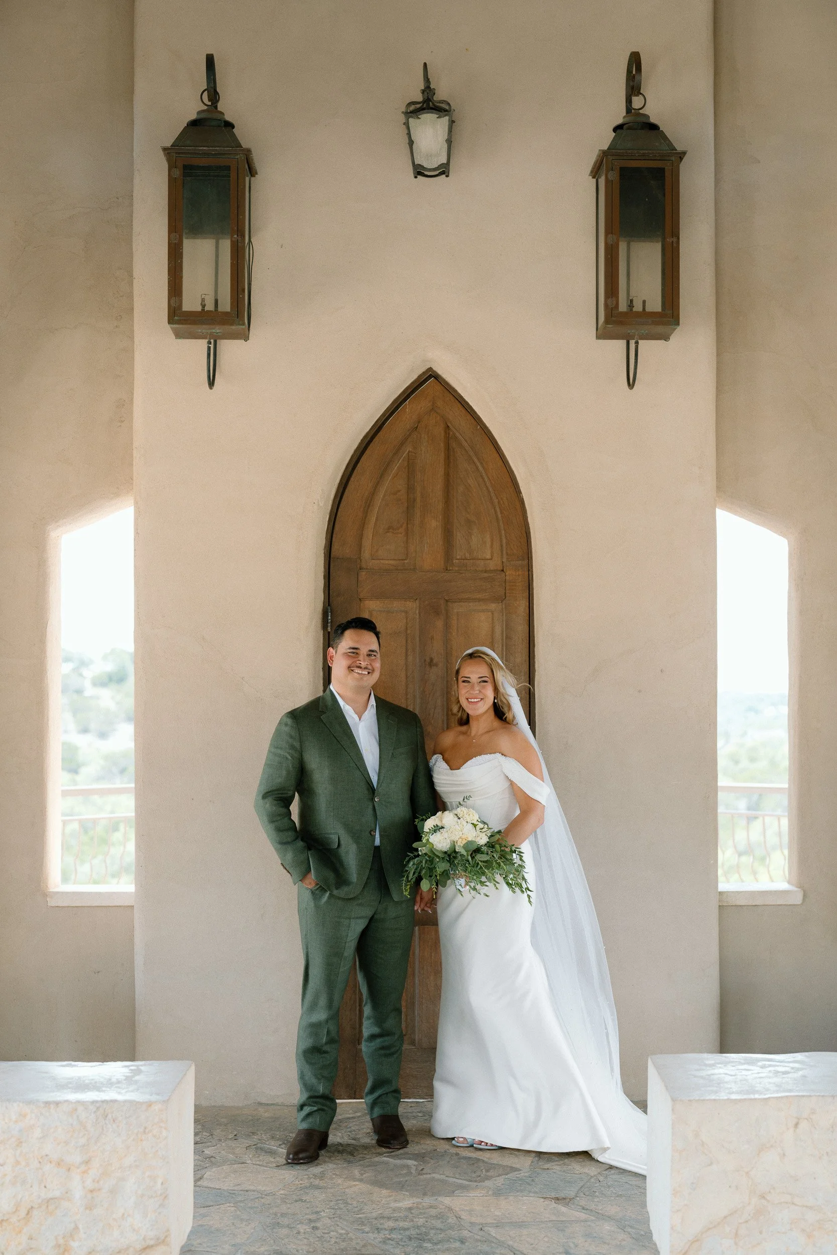 Bride and groom smile for a portrait in front of the arched wooden door at Chapel Dulcinea