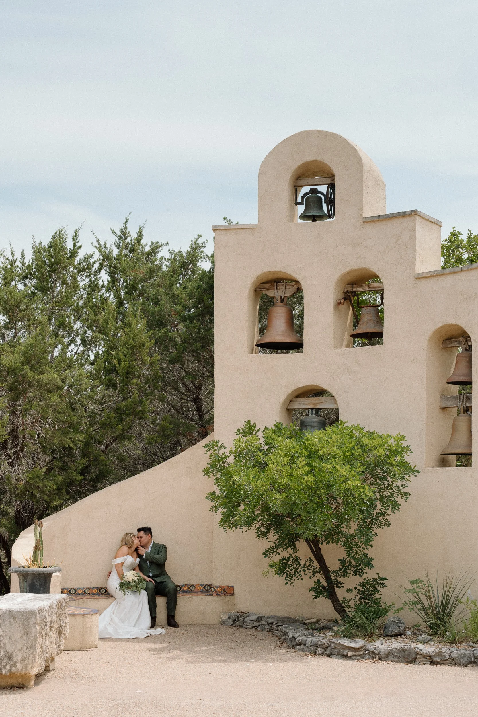 The newlyweds kiss on a tiled bench as the mission bell tower rises behind them at Chapel Dulcinea