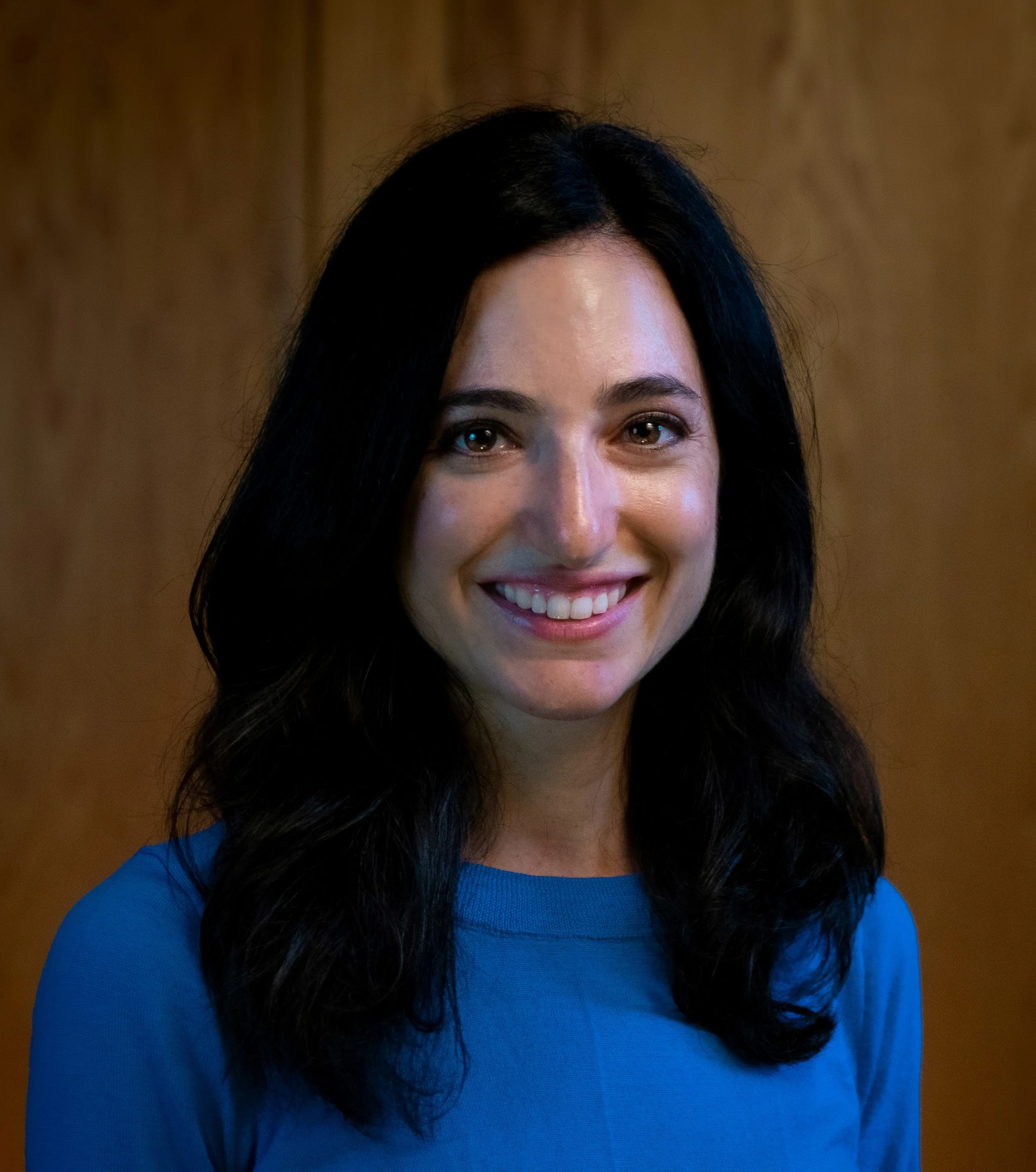 A woman with long black hair wearing a blue top, smiling, in front of a wooden background.