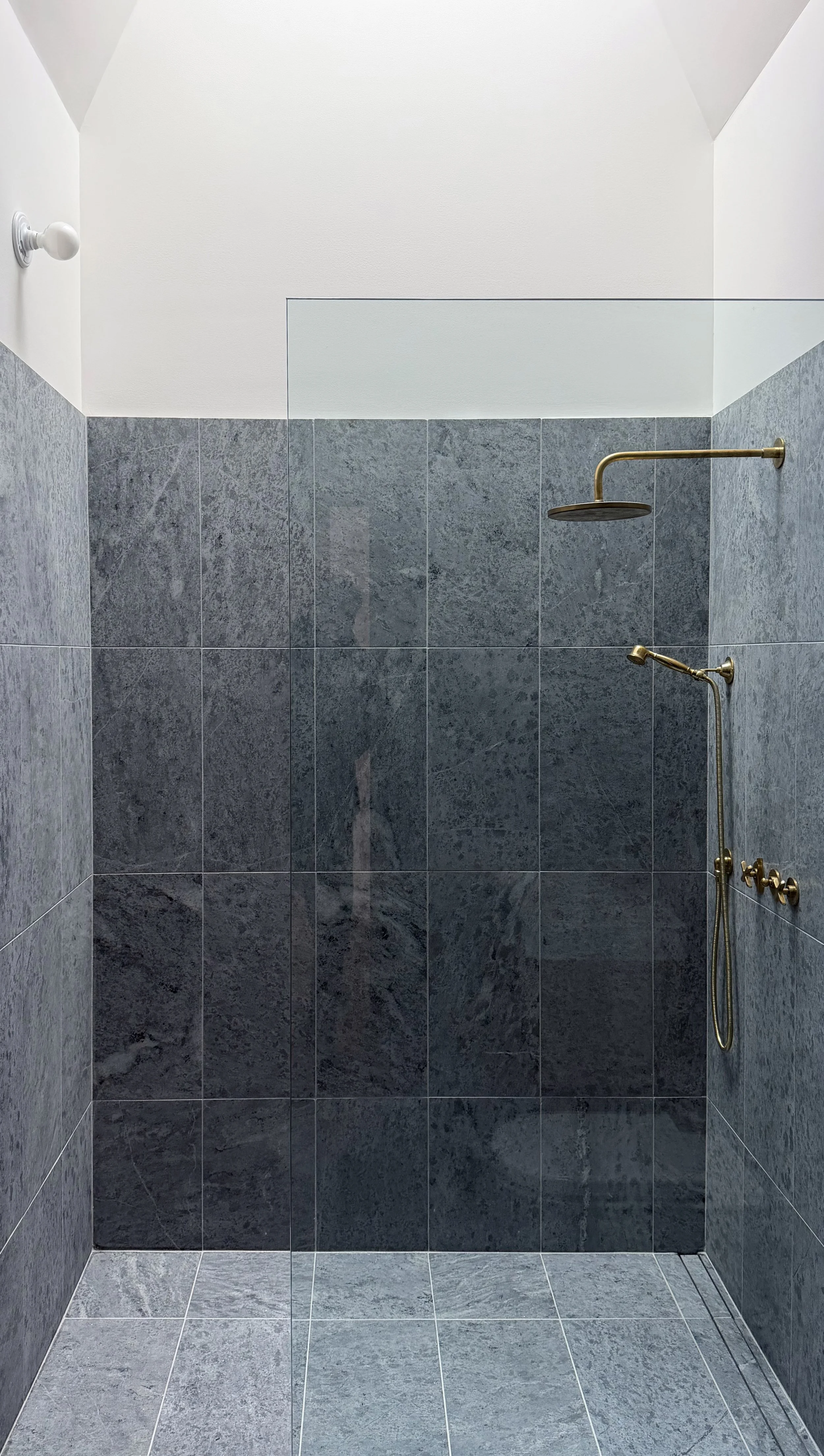 Shower area with gray tiled walls and floor, brass showerhead, handheld shower, and faucet.
