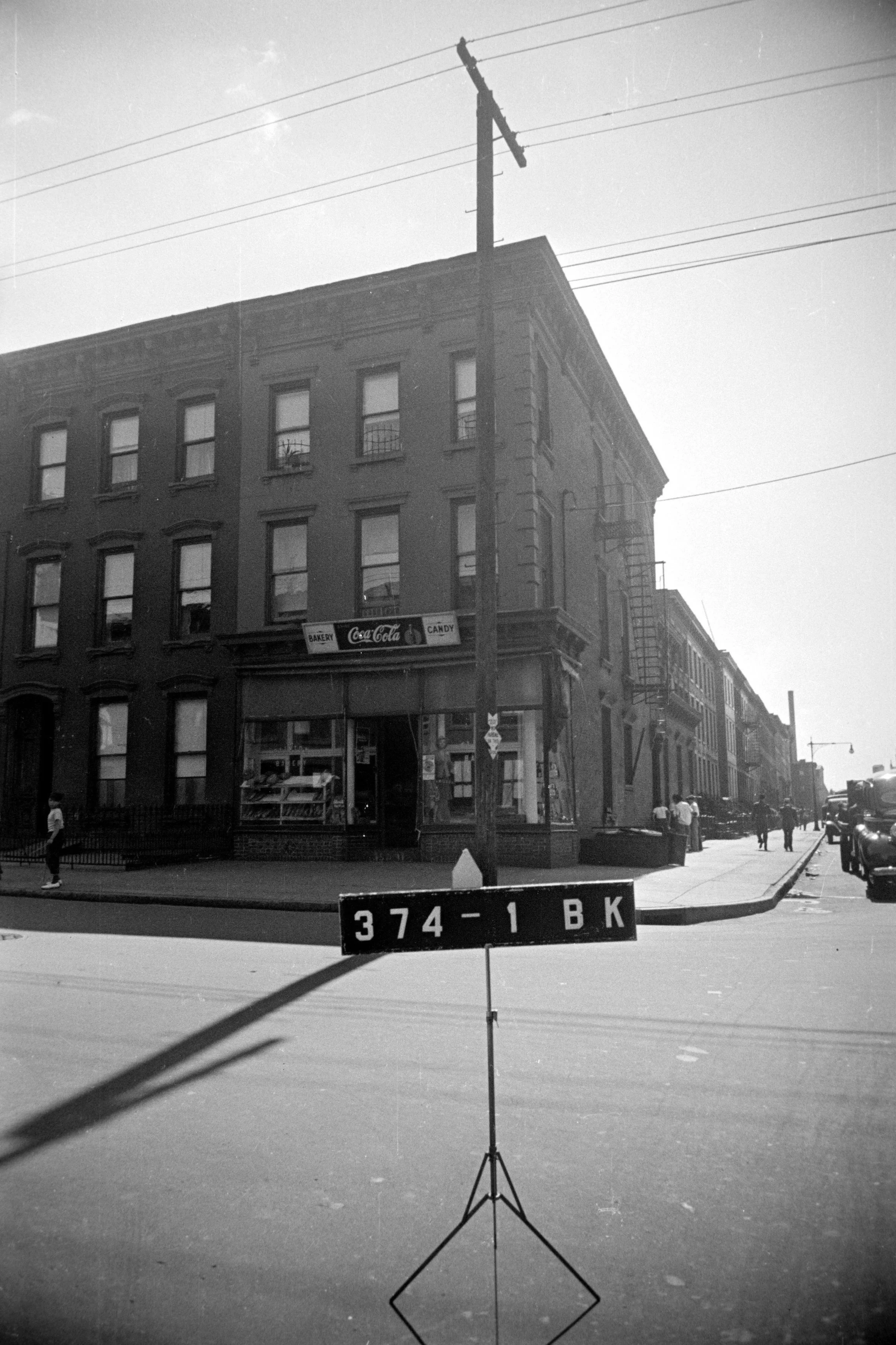 Black and white photo of a city street corner with a building that has a bakery, Coca-Cola, and candy signs, a fire escape, and a utility pole in the foreground with a road sign reading '374-1 BK'.