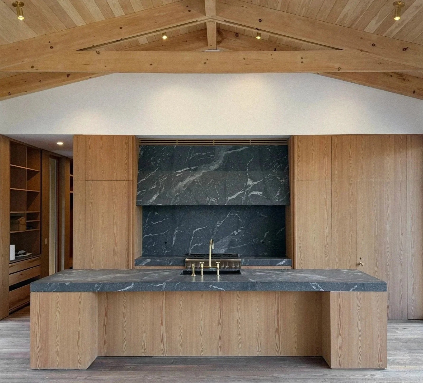 Modern kitchen with a large wooden island, dark marble countertop, and matching dark marble backsplash, wooden cabinetry, and a vaulted ceiling with exposed wooden beams.