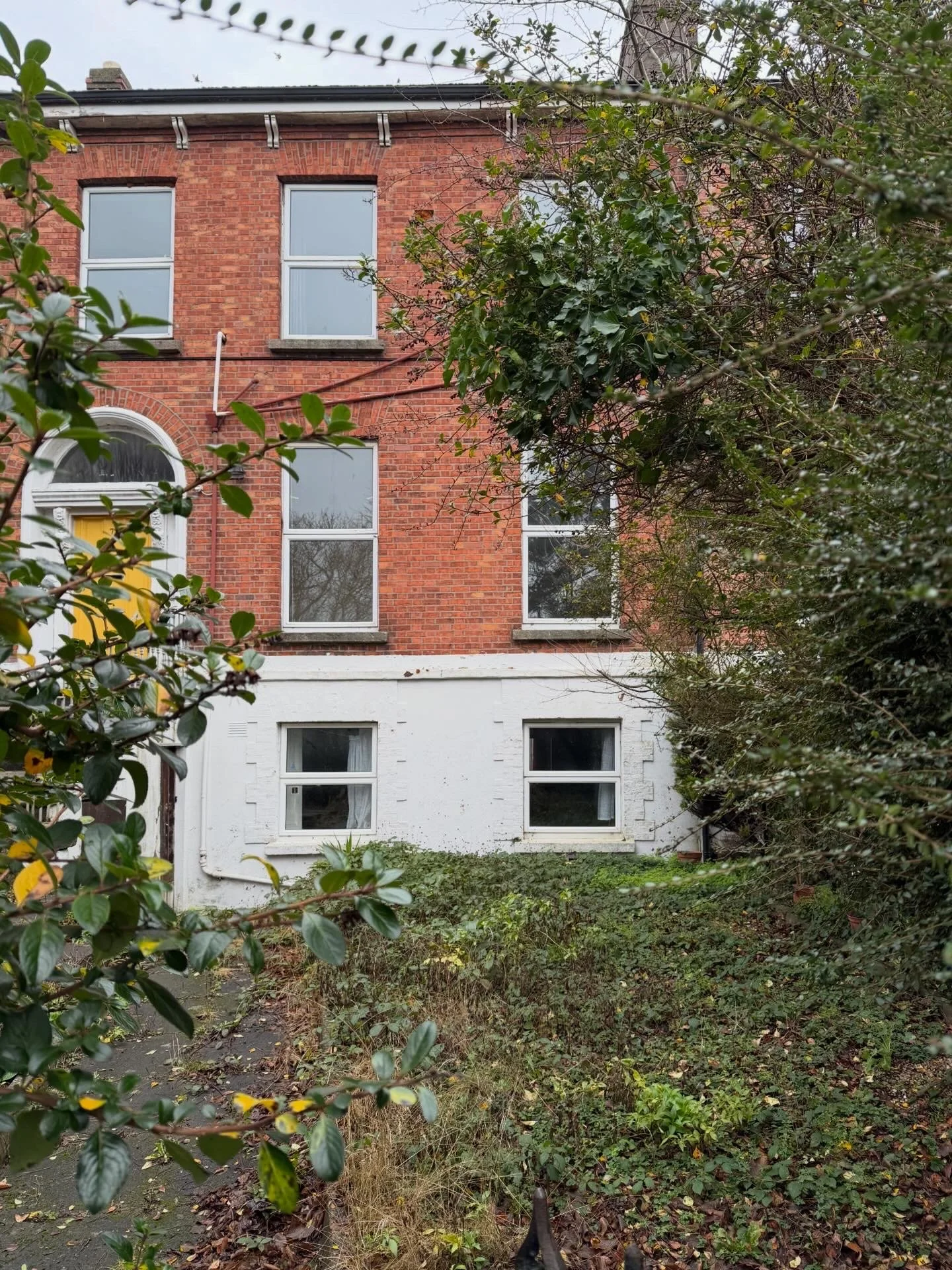 Front view of a four-story brick building with white window frames, partially obscured by trees and bushes in the foreground.