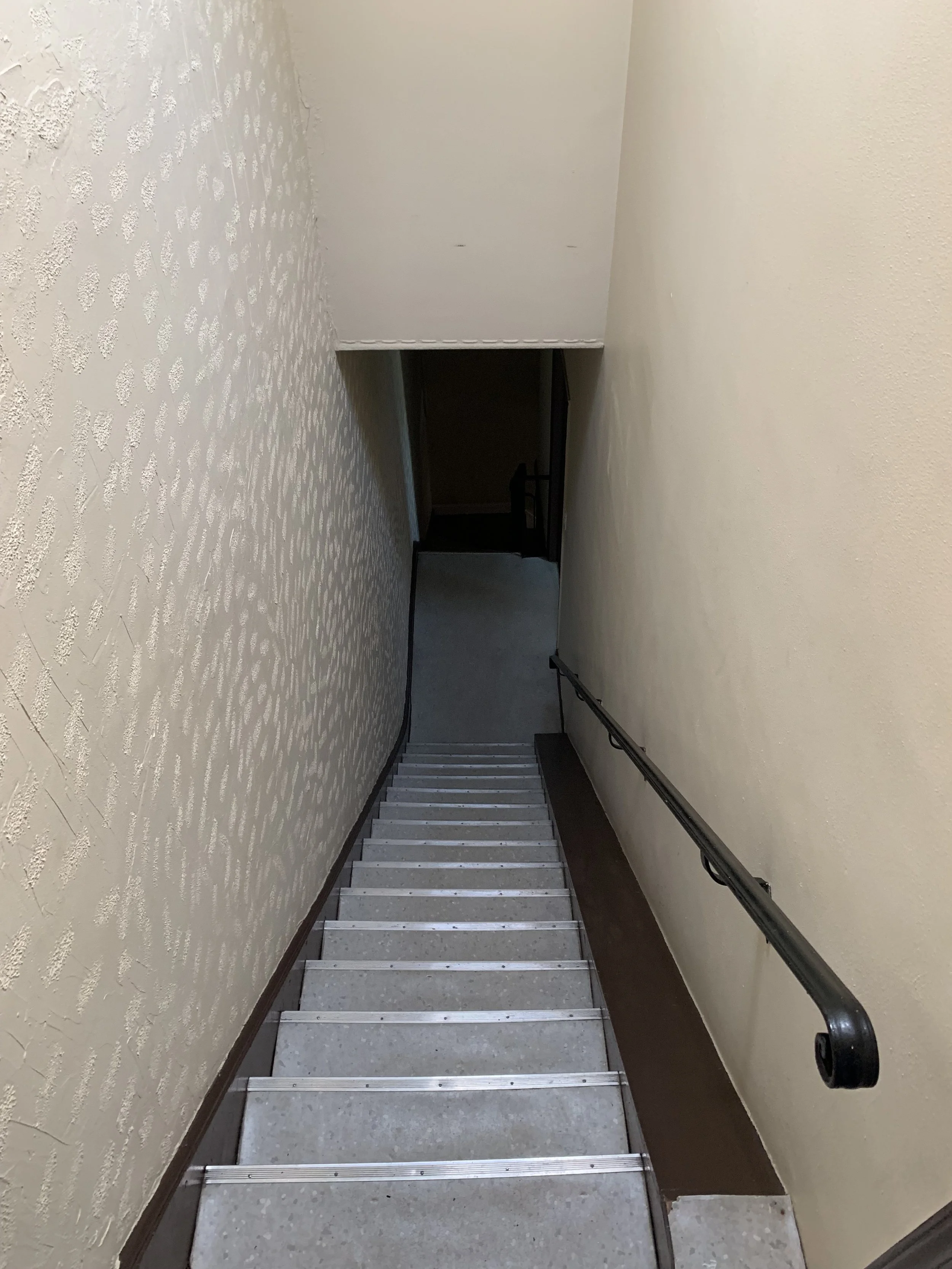 A staircase seen from the top, descending into a dark hallway with a handrail on the right side and textured wall on the left.
