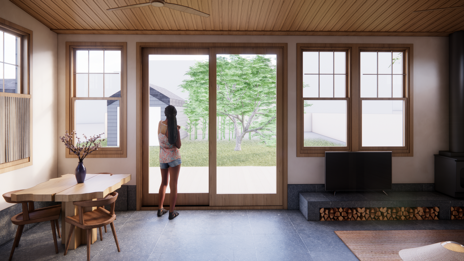 A woman standing inside a room with wooden-paneled ceiling and large windows, looking out at a backyard with grass, a tree, and white walls.