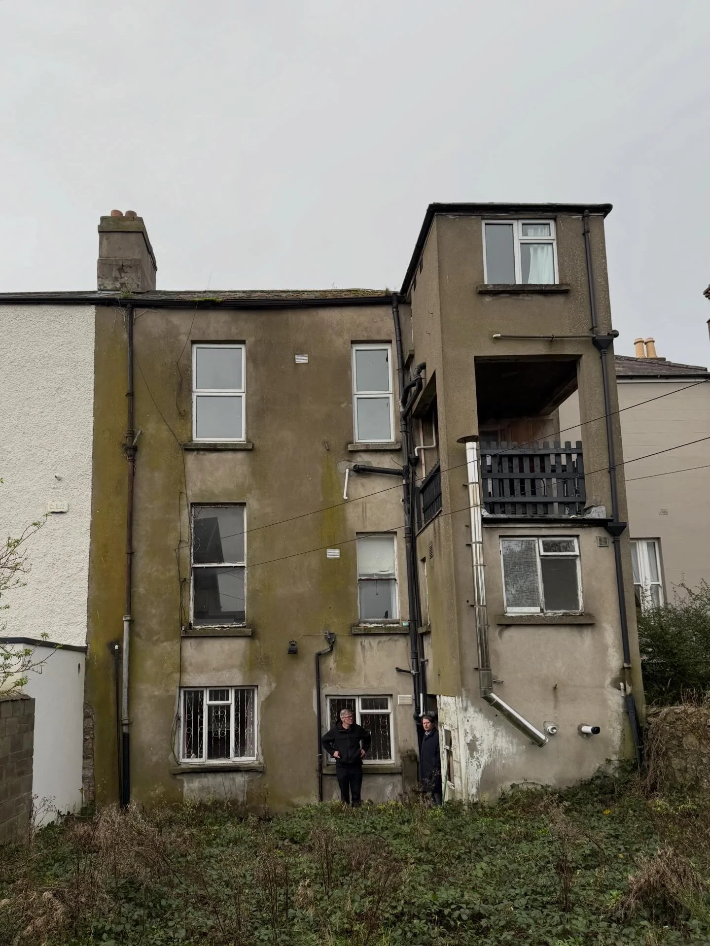 An old, weathered apartment building with moss and dirt on its facade, two men standing in front, overgrown yard, cloudy sky.