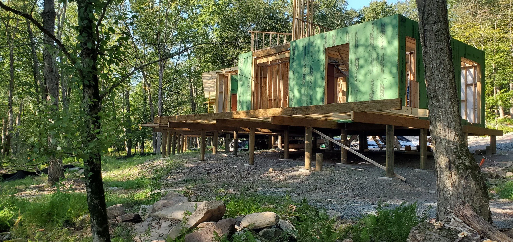 Under construction house framed with green sheathing in a wooded area, on stilts with outdoor staircase, surrounded by trees and rocks.