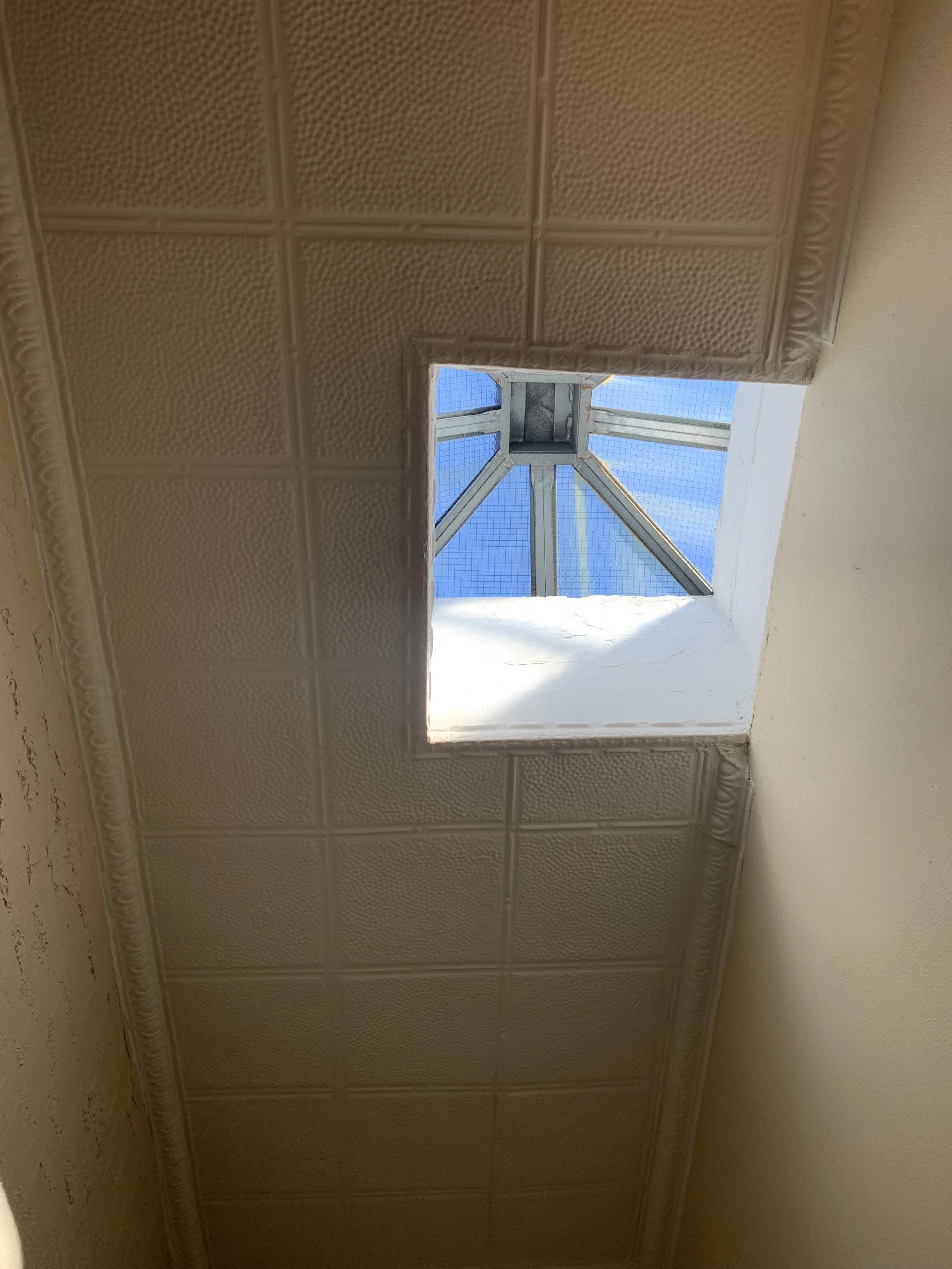 Looking up at a small square skylight in the ceiling, showing part of the sky and a metal frame with support beams.