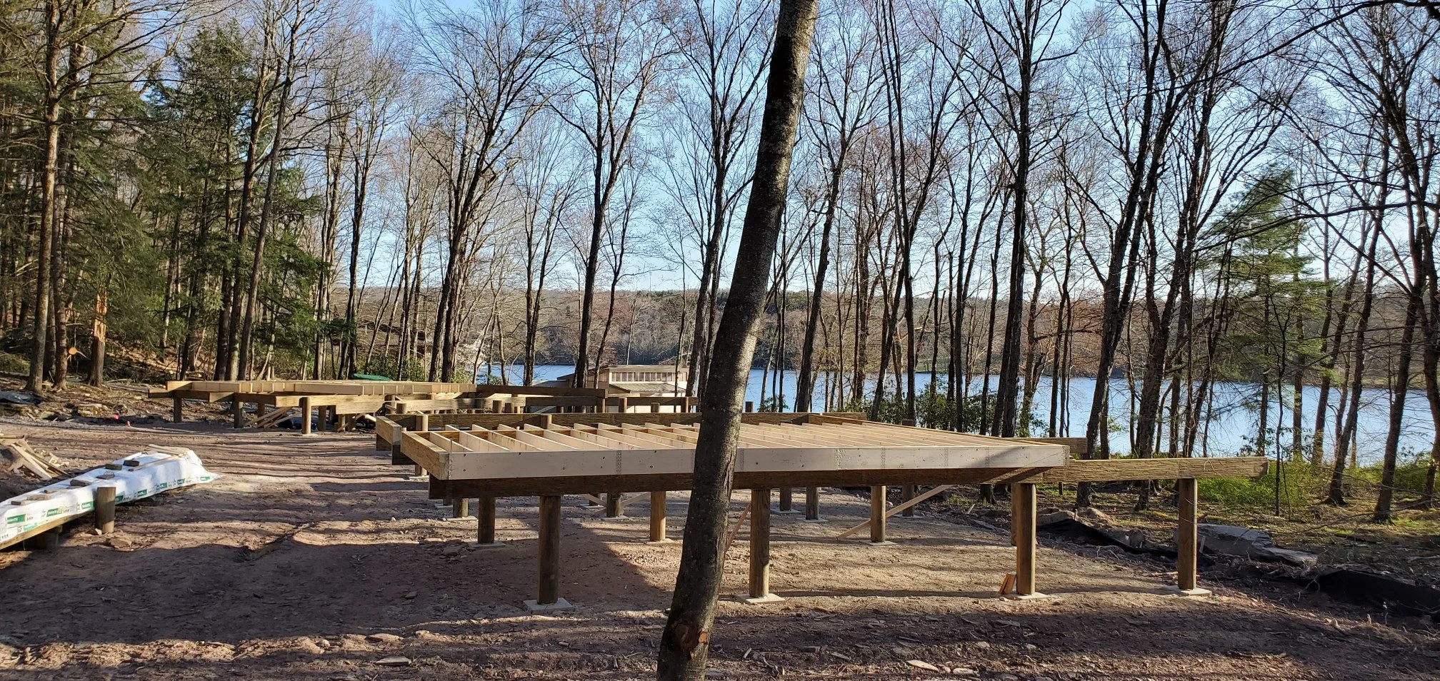Construction site in progress with wooden framing for a deck overlooking a lake, surrounded by trees and a clear sky.