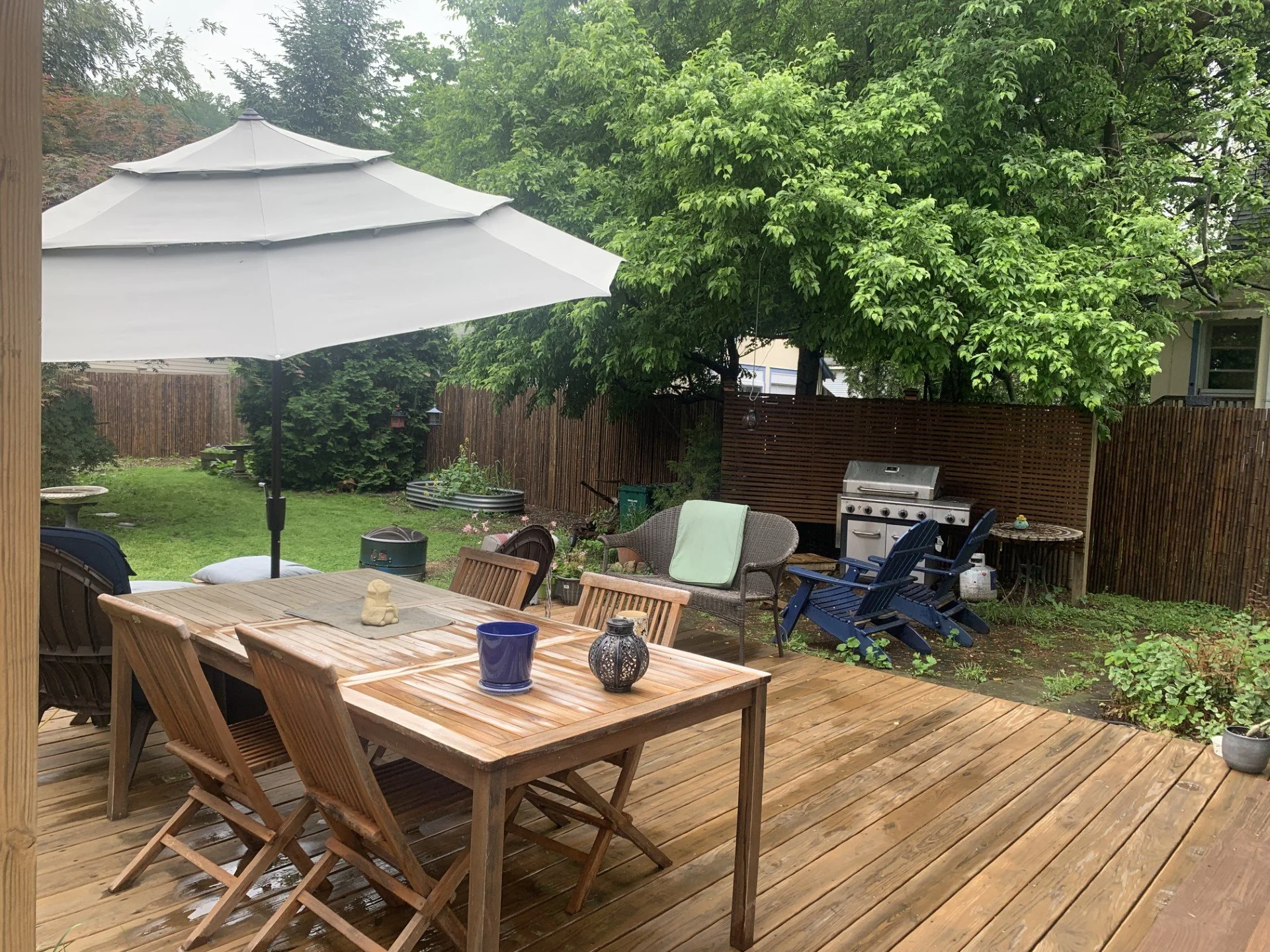 Patio scene with a large wooden table, six chairs, a blue umbrella, a granite-top grill, a small round table, a garden with green trees, potted plants, and a wooden fence.