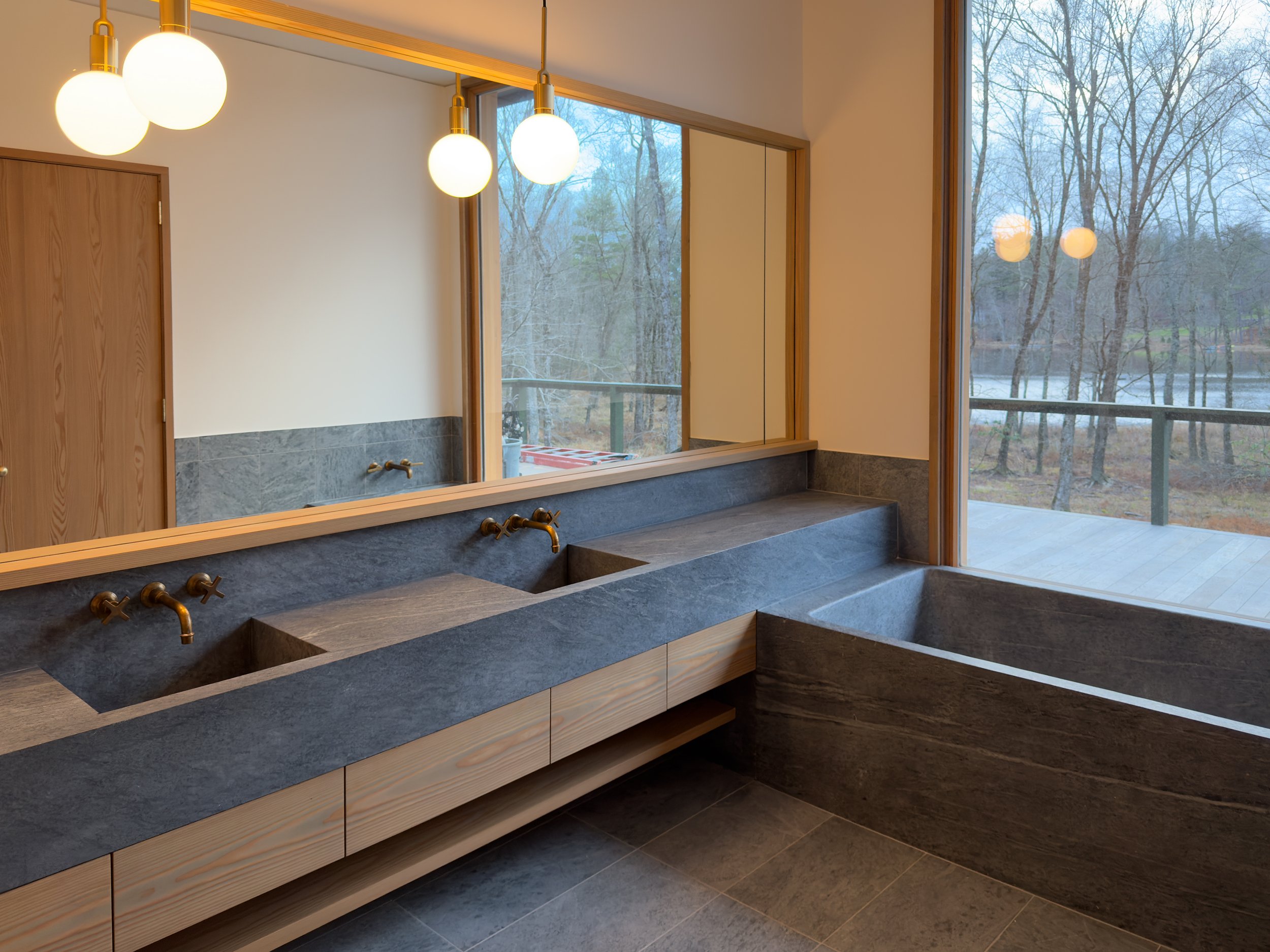 Modern bathroom with dark stone sink and bathtub, large window showing outdoor trees and lake, pendant light fixtures, wooden accents.