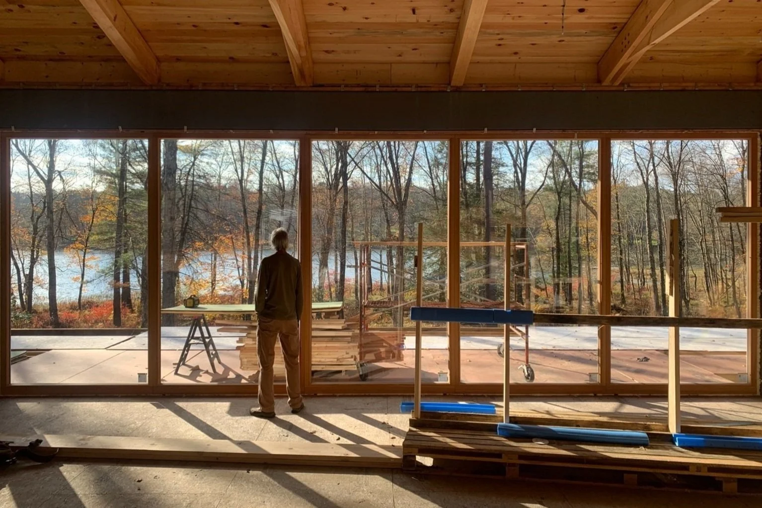 A person standing inside a house under construction, looking out through large floor-to-ceiling windows at a wooded landscape with trees and a lake.