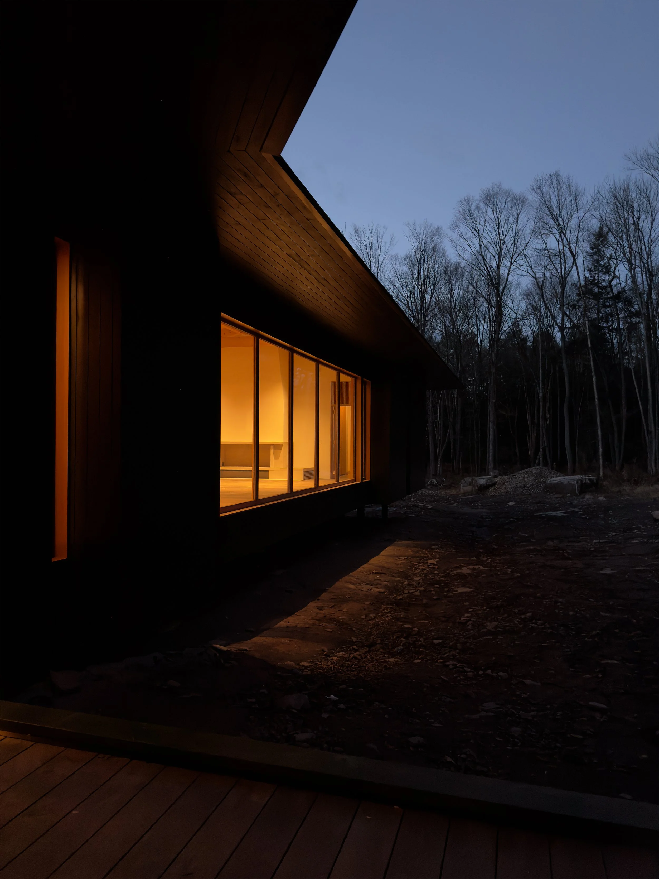 Modern house at dusk with warm interior lighting, large glass windows, and surrounded by leafless trees.