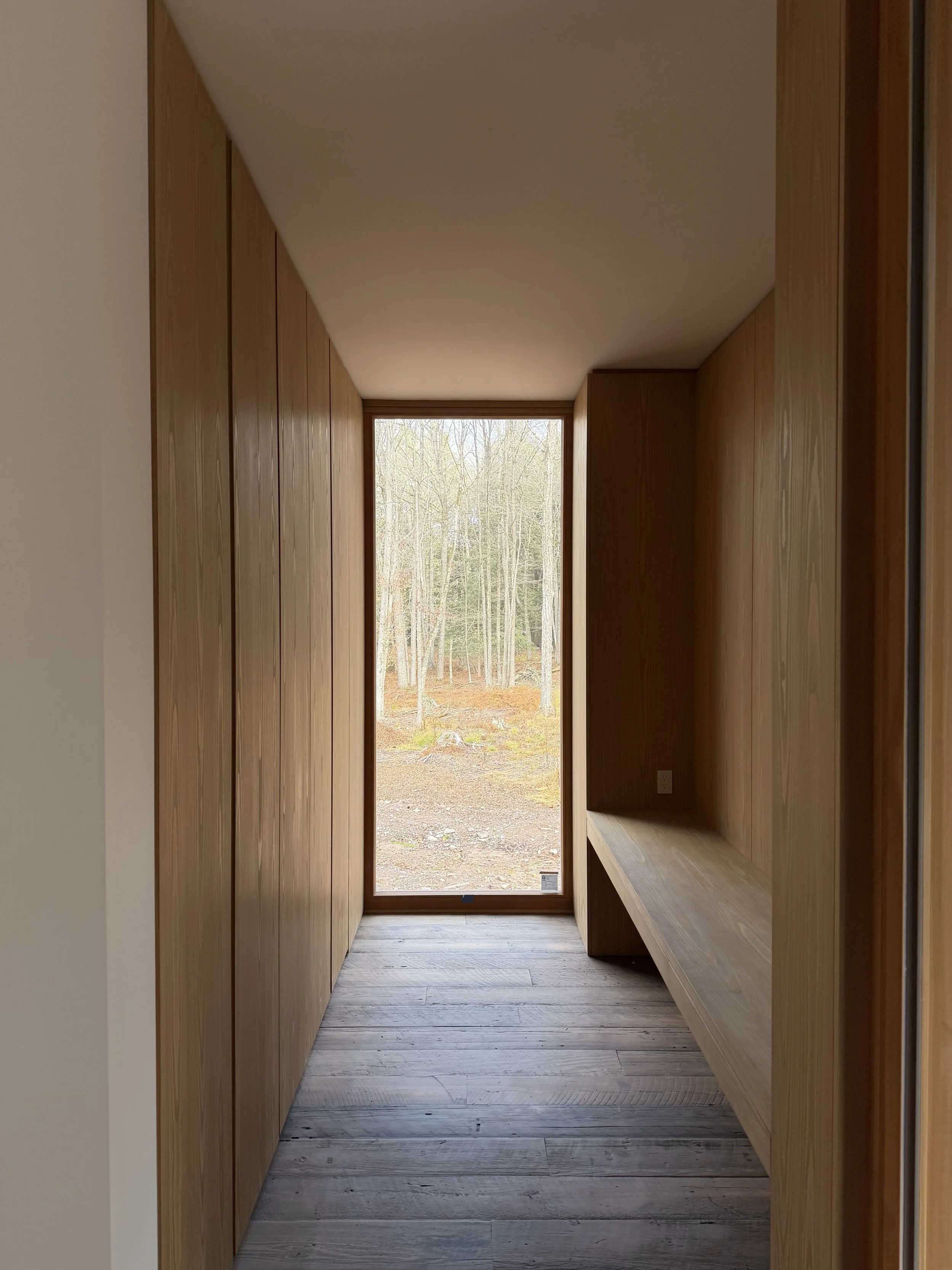 Narrow hallway with wooden walls and a large window at the end showing a forest outside, with a wooden bench on the right side.