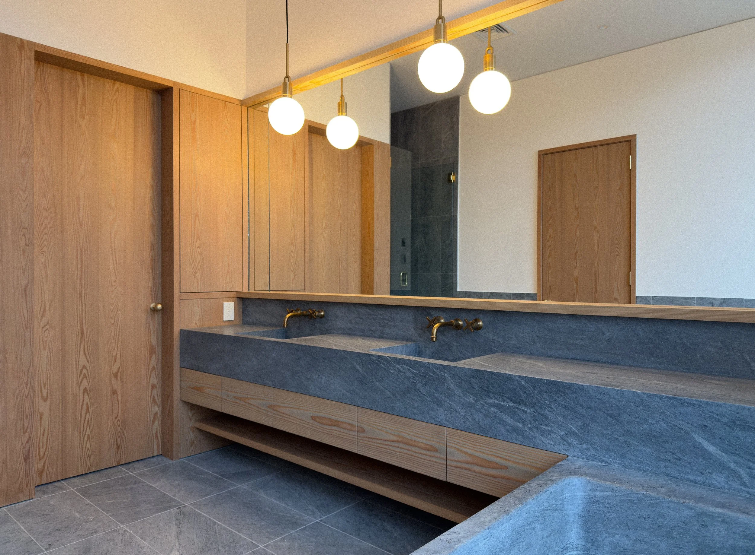 Modern bathroom with a large mirror, double sink with gold faucets, wooden cabinets, and a stone countertop, illuminated by pendant lights.