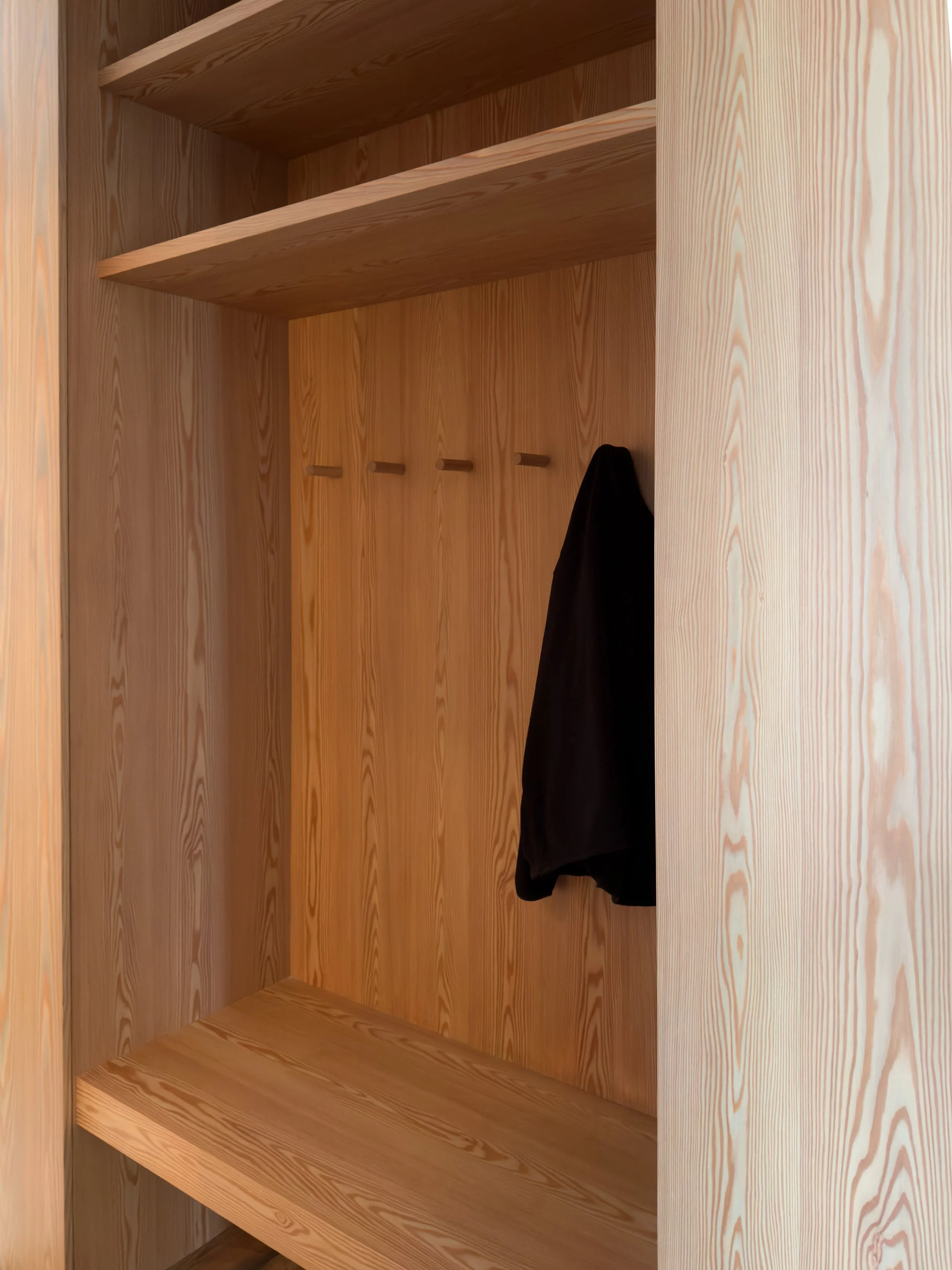 Empty wooden closet with a single black jacket hanging and three wooden hooks.