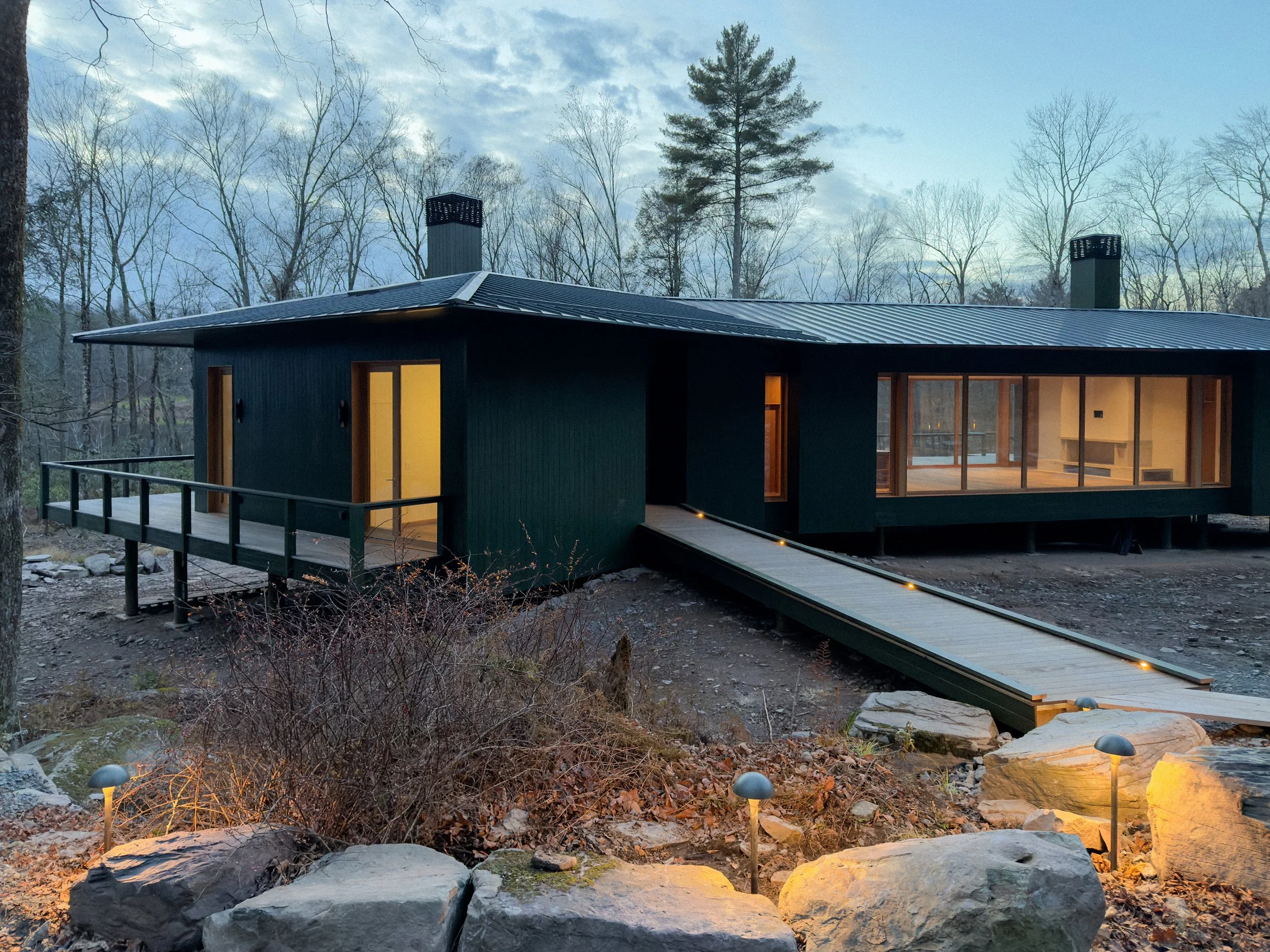 Modern black house with large glass windows, a wooden ramp with lights leading to the entrance, surrounded by rocks and trees, under a blue sky with scattered clouds.
