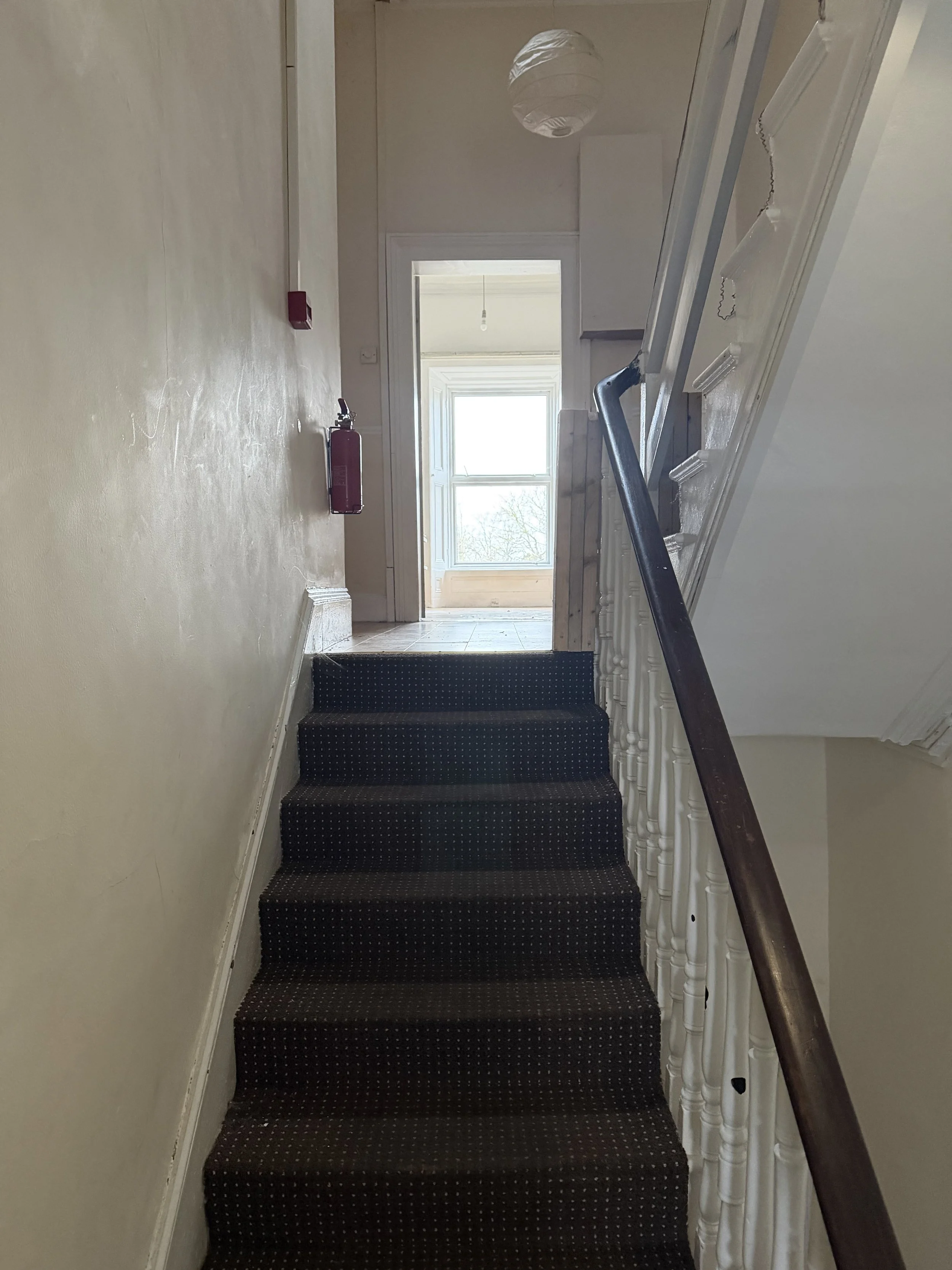 A staircase with a dark patterned carpet, white banister on the right, leading to a bright room with large windows at the top. There is a fire extinguisher mounted on the left wall and a ceiling light fixture.