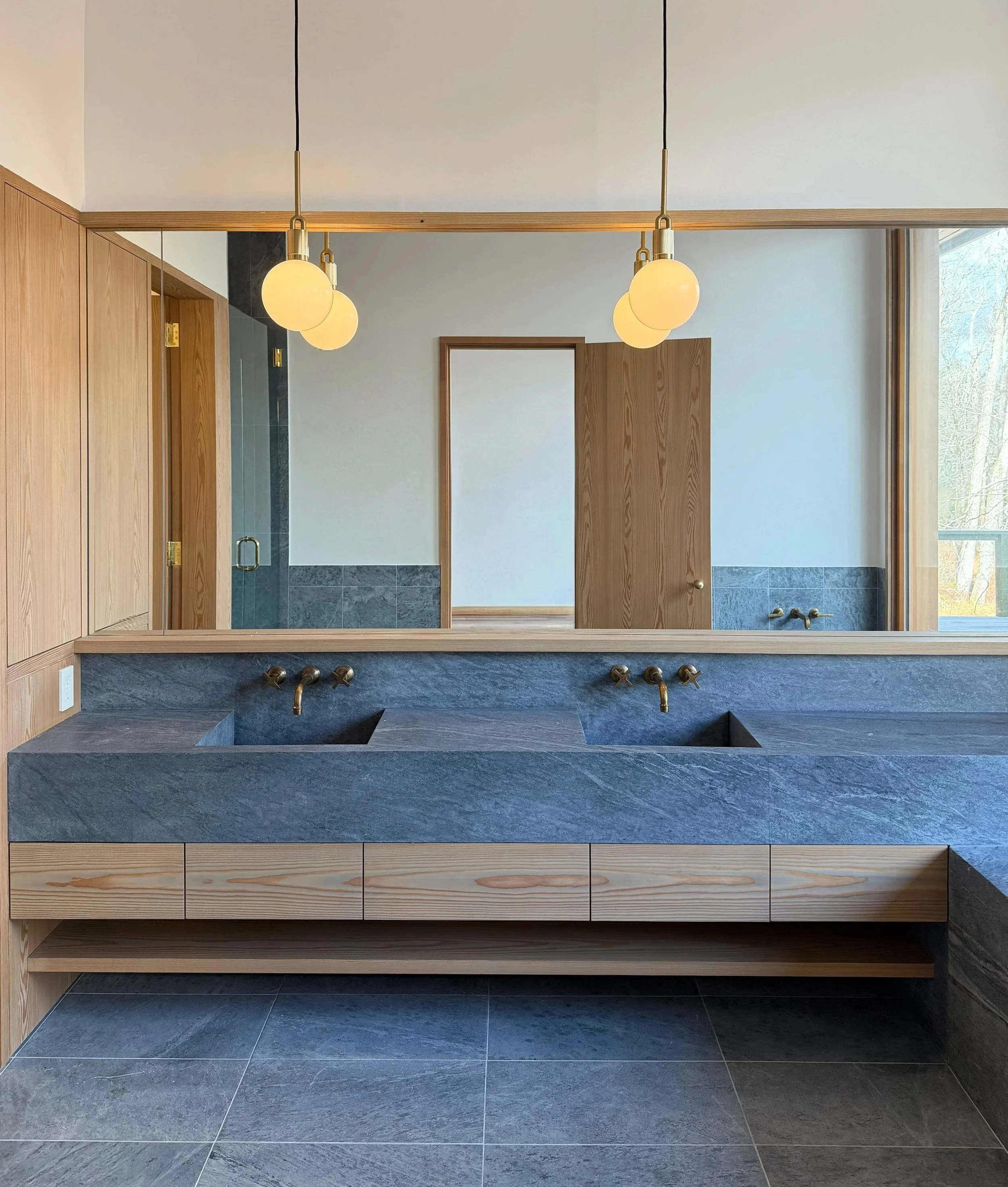 Modern bathroom with double sink vanity, dark gray stone countertop, brass fixtures, and a large mirror, with wooden accents and a window to the right.
