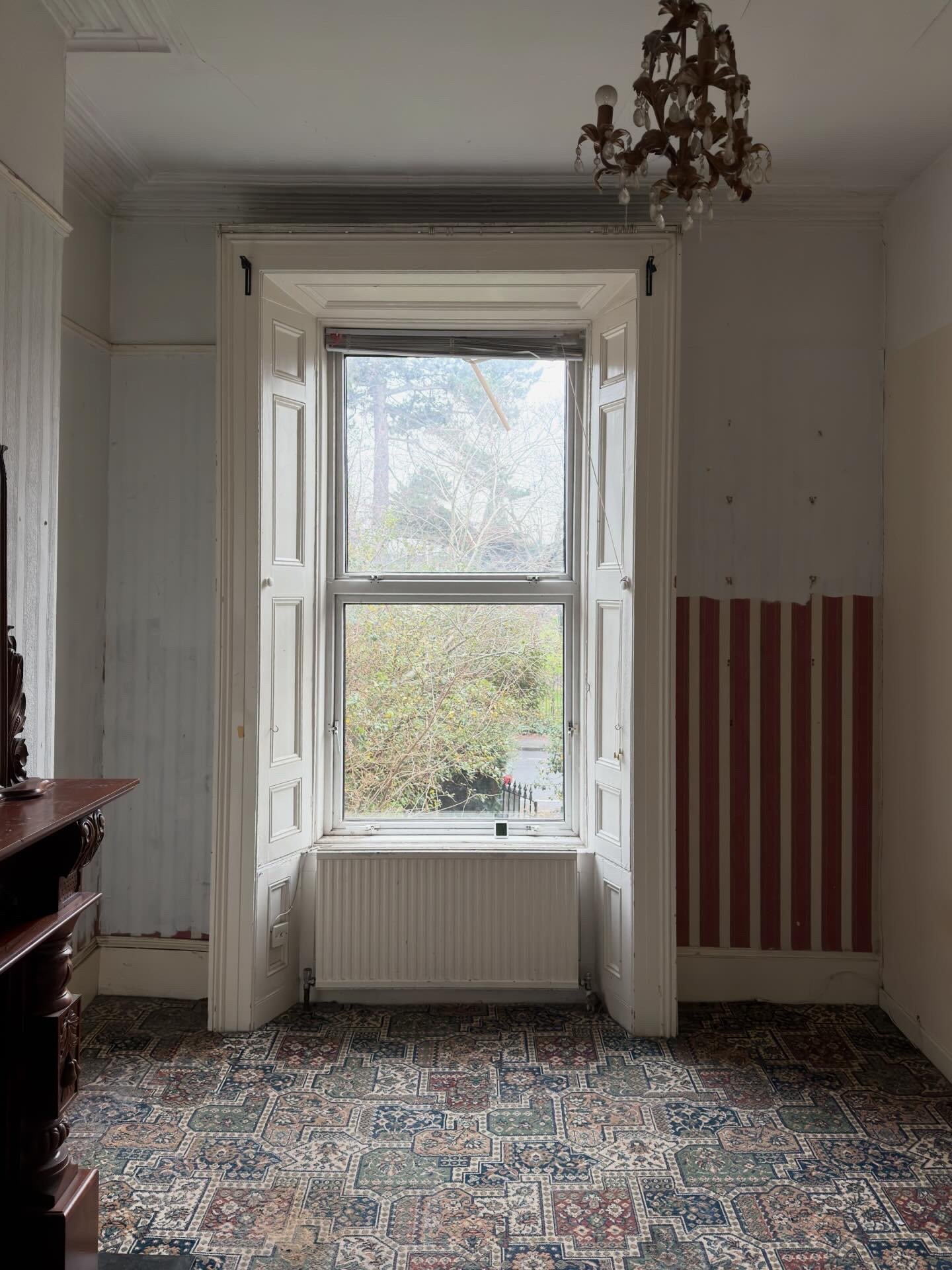 Room with a large window, chandelier hanging from the ceiling, patterned carpet on the floor, and a partially stripped wall with red and white vertical stripes.