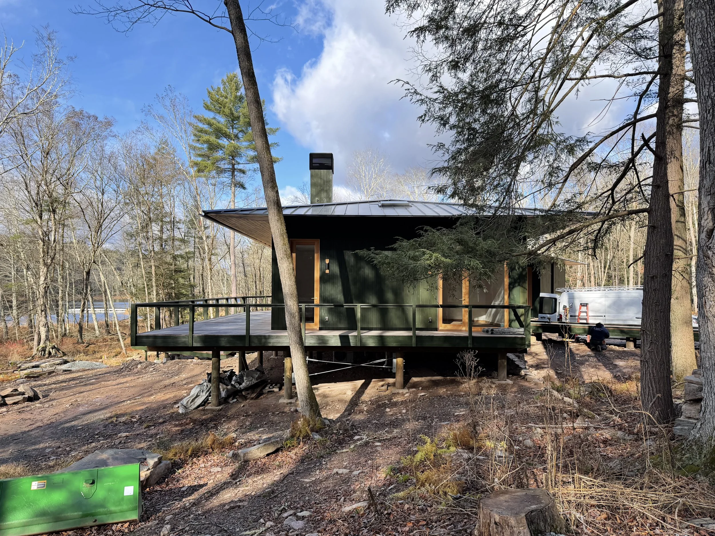 A modern house under construction in a forested area, with a deck, trees, and construction equipment visible.