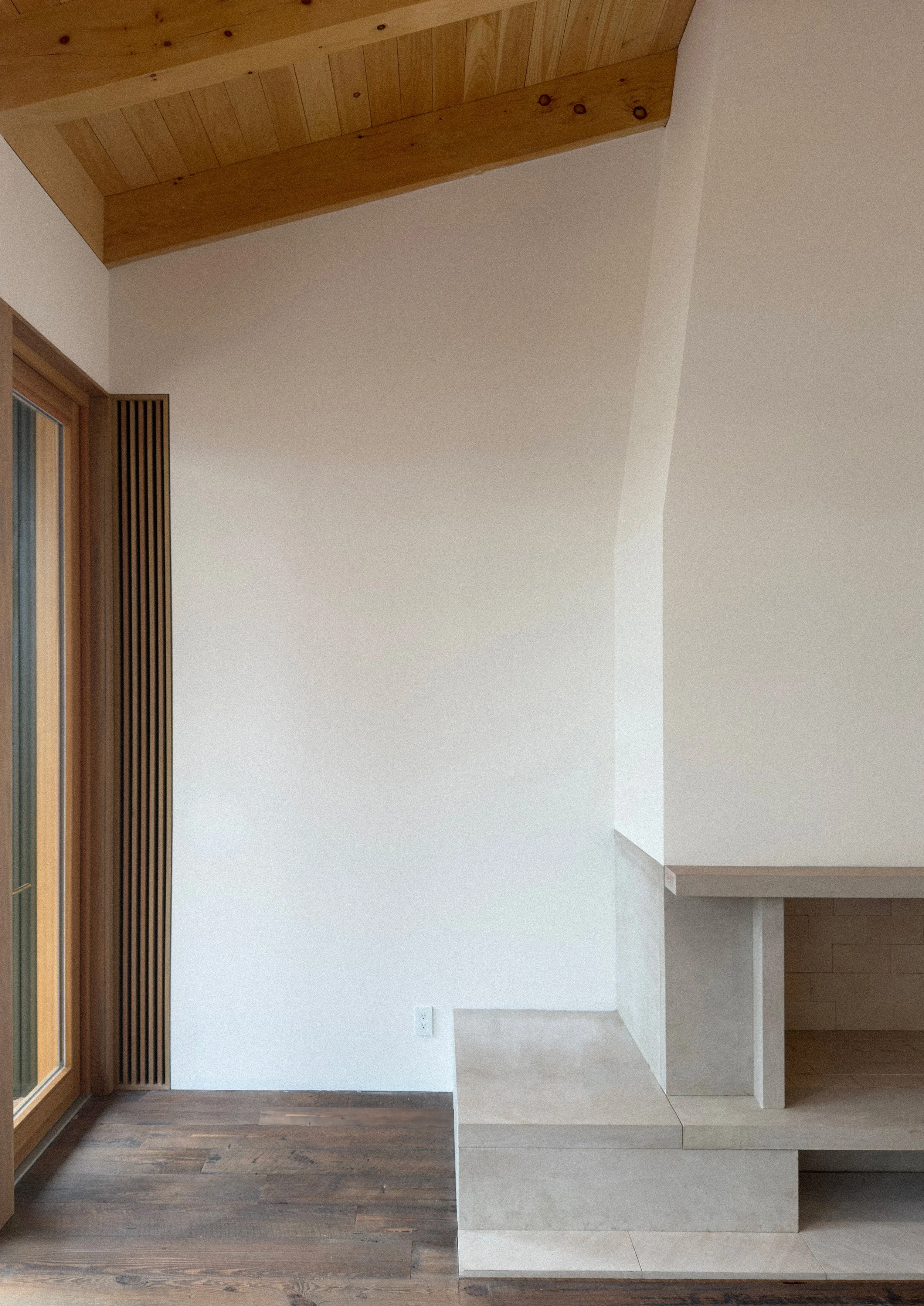 Interior of a modern, minimalist living space with wooden flooring, a white wall, a corner with concrete and wood design elements, and a door with wooden slats.