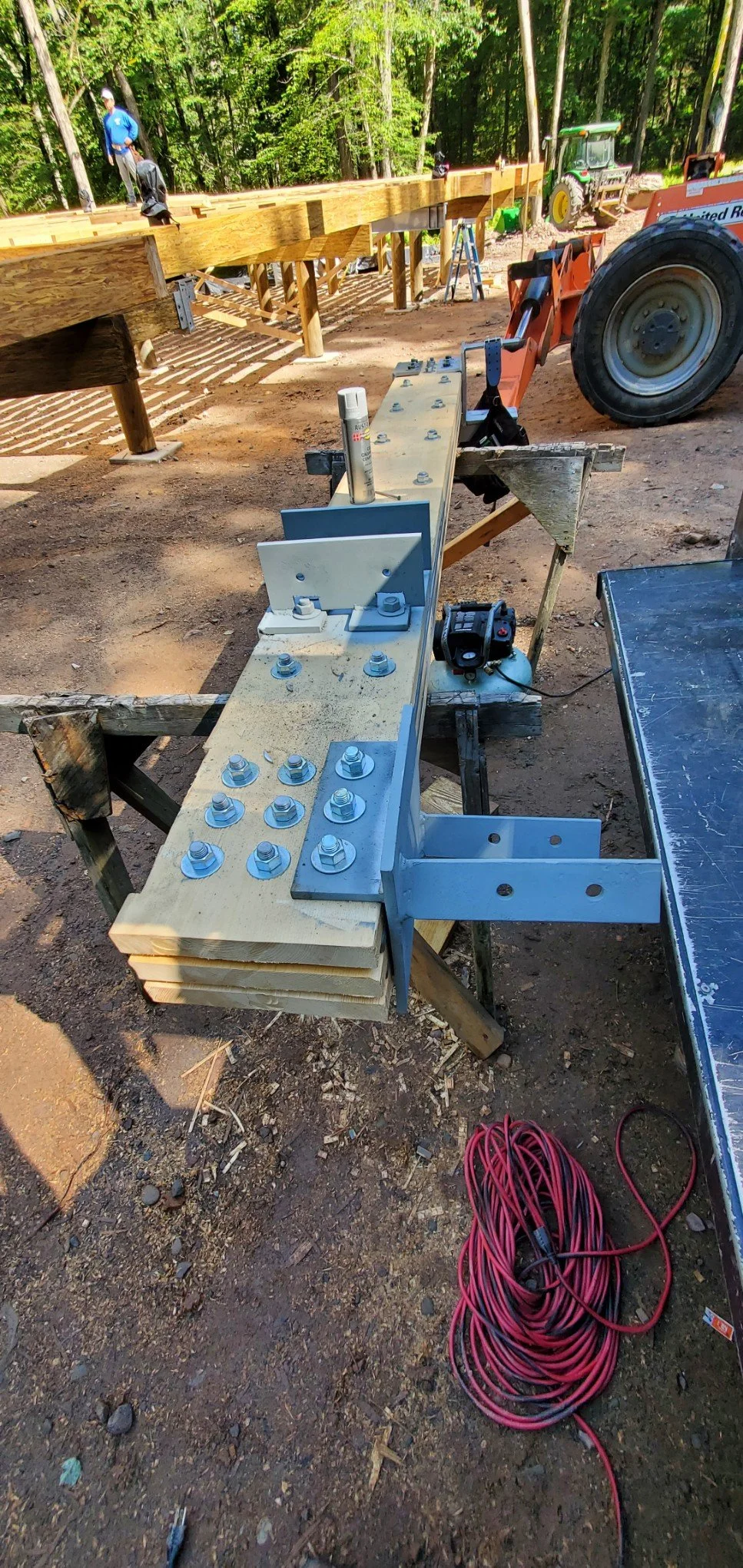 Construction site with wooden framework, machinery, and workers assembling a bridge or similar structure in a wooded area.