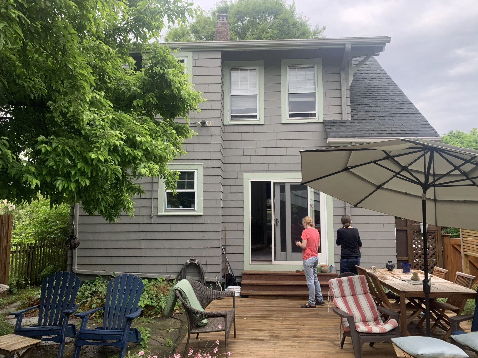 A backyard patio with wooden deck, outdoor furniture, large umbrella, and two women standing near the house, which has gray siding and multiple windows. A tree with green leaves is on the left side of the image.