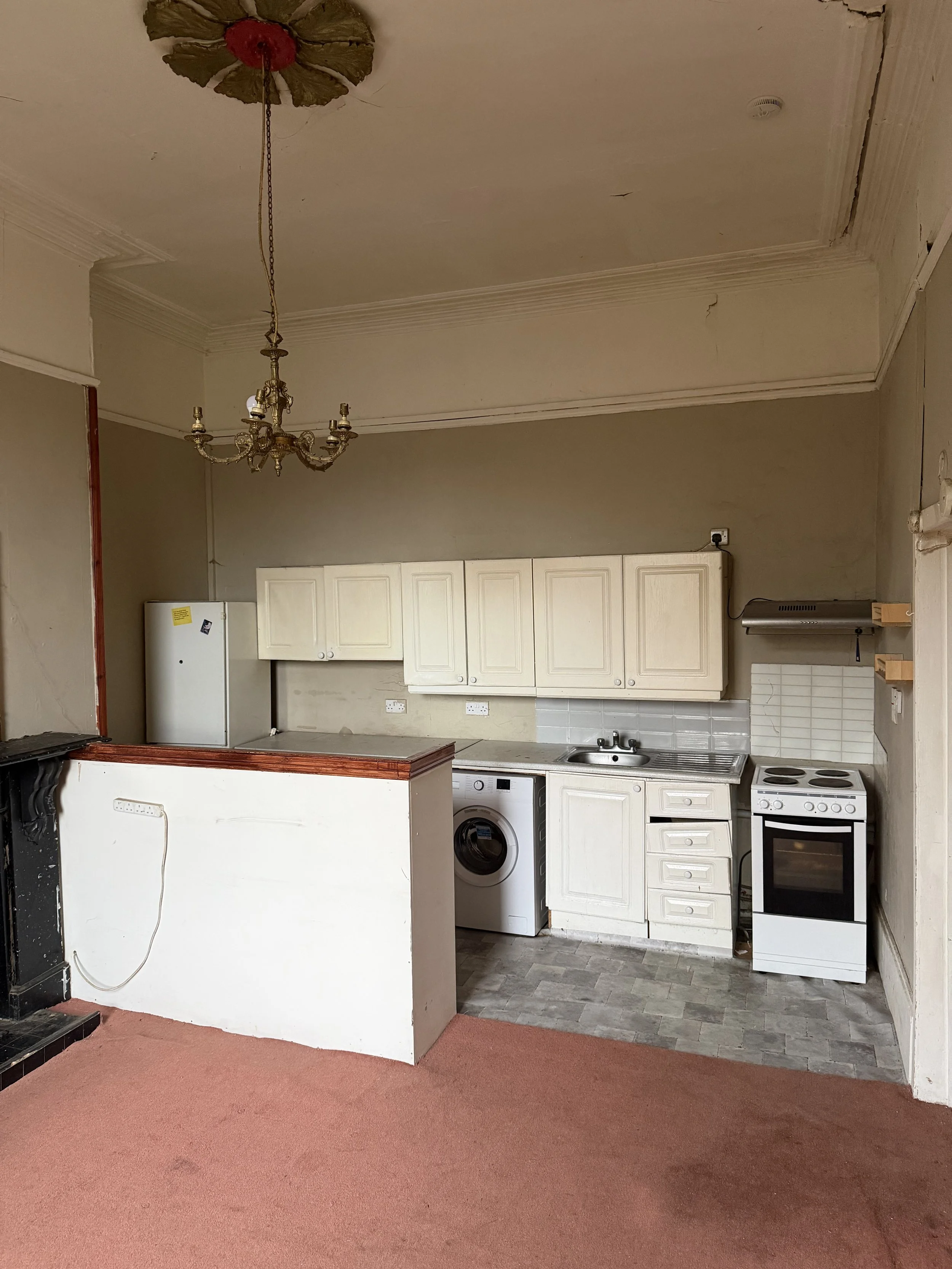 Small kitchen area with white cabinets, a washing machine, a stove, and a sink, with a chandelier hanging from an outdated ceiling and worn wallpaper.