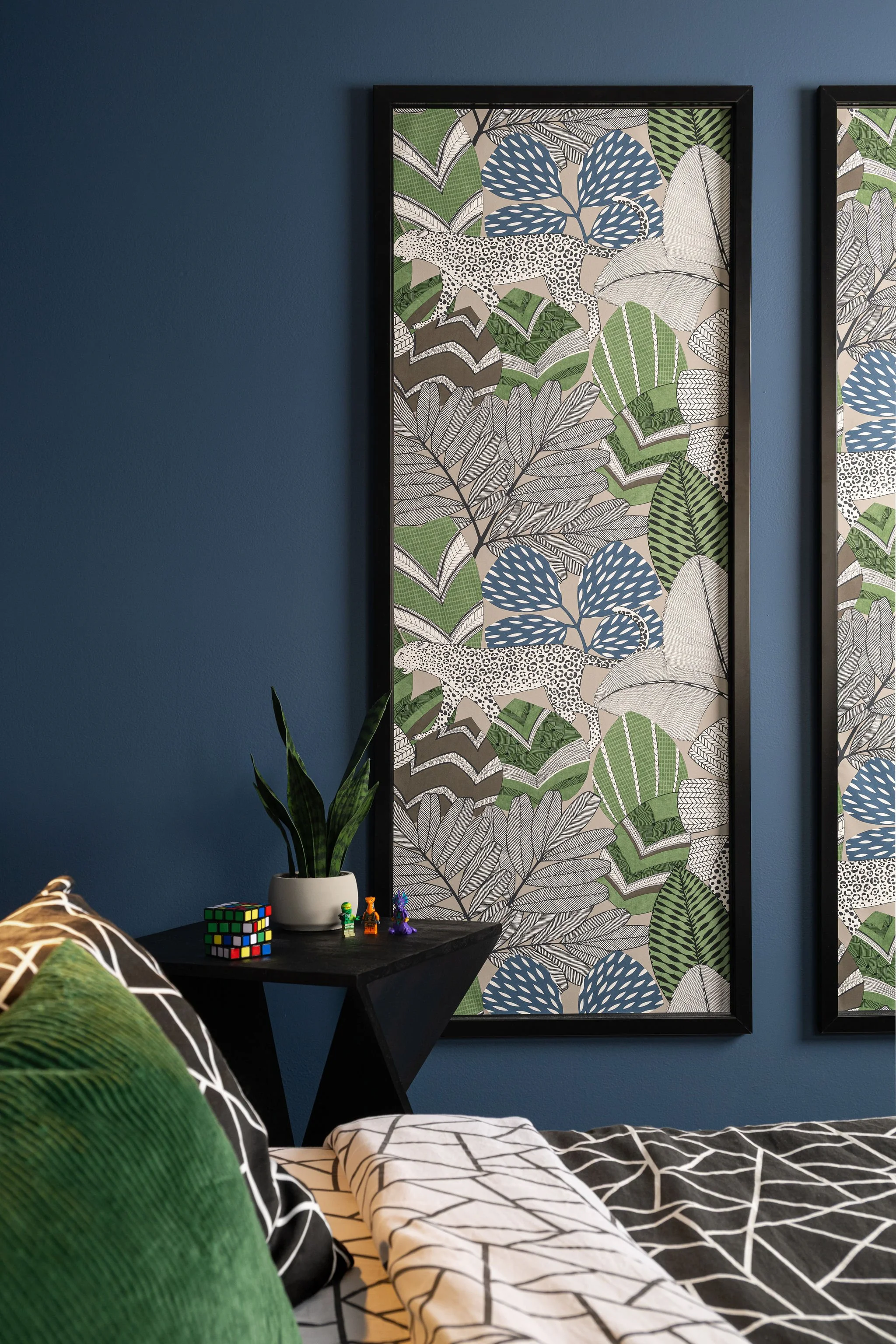 A bedroom with a blue wall featuring large botanical wallpaper with leaves, cheetahs, and abstract patterns framed in black. A black nightstand holds a plant in a white pot, a Rubik's cube, and small figurines. The bed has black and white geometric bedding and a green pillow.
