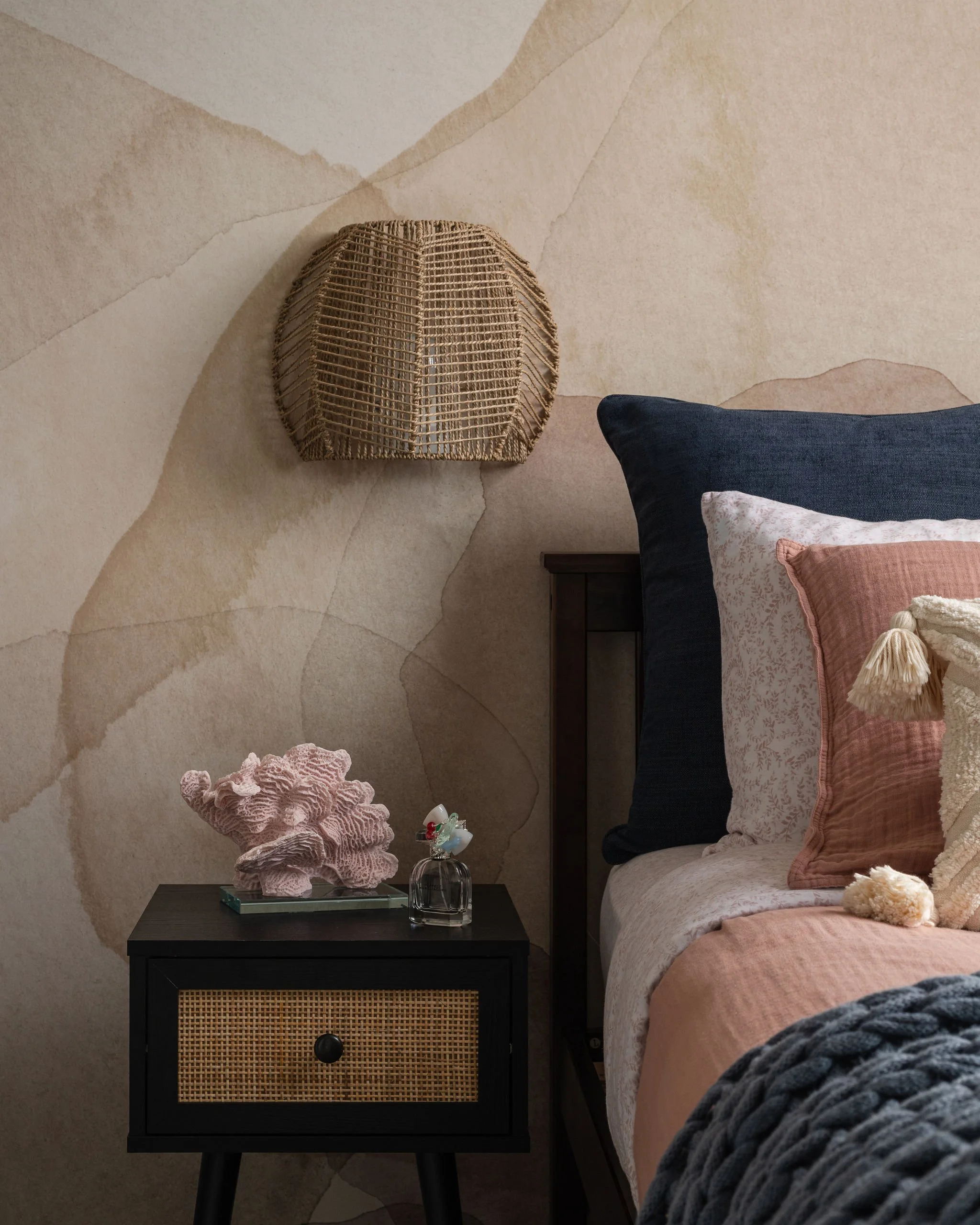 A bedroom nightstand with a glass bottle, a decorative coral piece, a book, and a cozy blanket, next to a bed with purple, pink, and patterned pillows and a white quilted blanket, against a beige artistically painted wall.