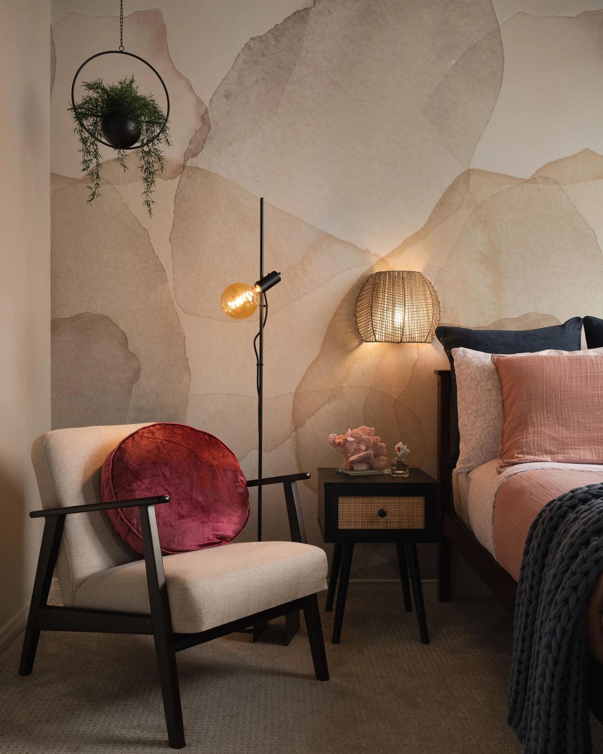 Cozy reading corner in a teen bedroom with upholstered chair, coral velvet pillow, floor lamp, hanging planter, woven wall sconce, blush bedding, and abstract mural wallcovering.