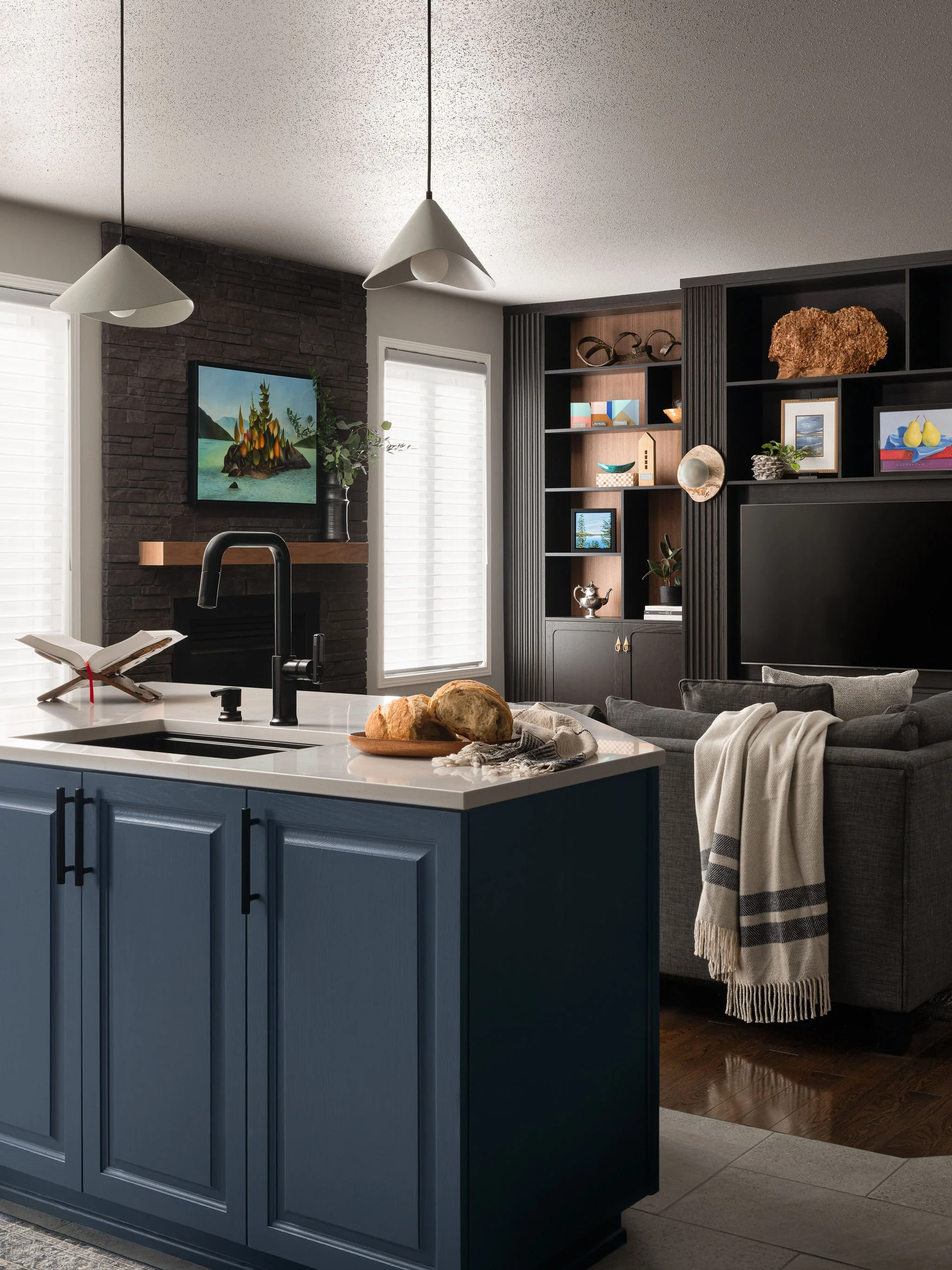Open-concept kitchen and living room with blue cabinetry, white quartz island, black faucet, dark stone fireplace, and custom black built-in media wall with styled open shelving.