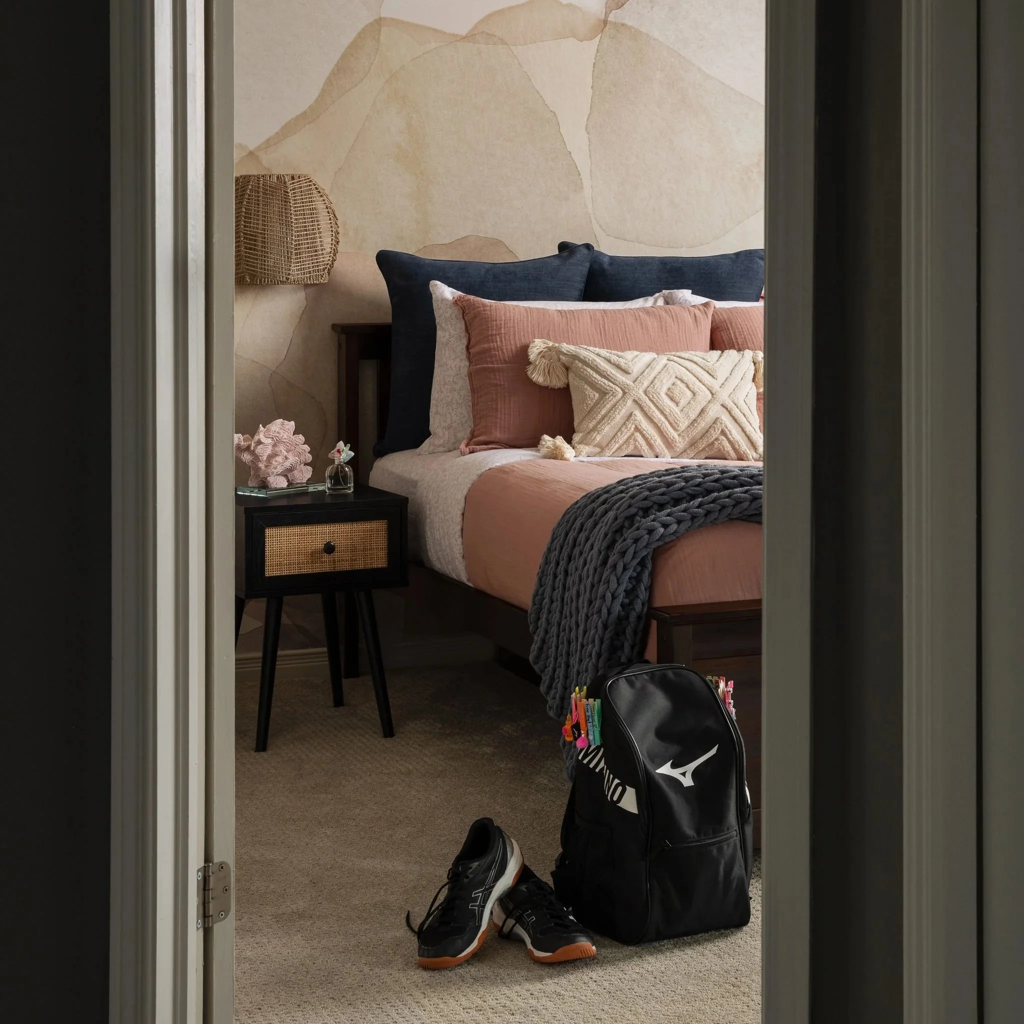 Teen bedroom viewed through an open doorway, featuring blush and coral bedding, a soft abstract mural wallcovering, woven wall sconce, cane nightstand, and school bag by the bed.