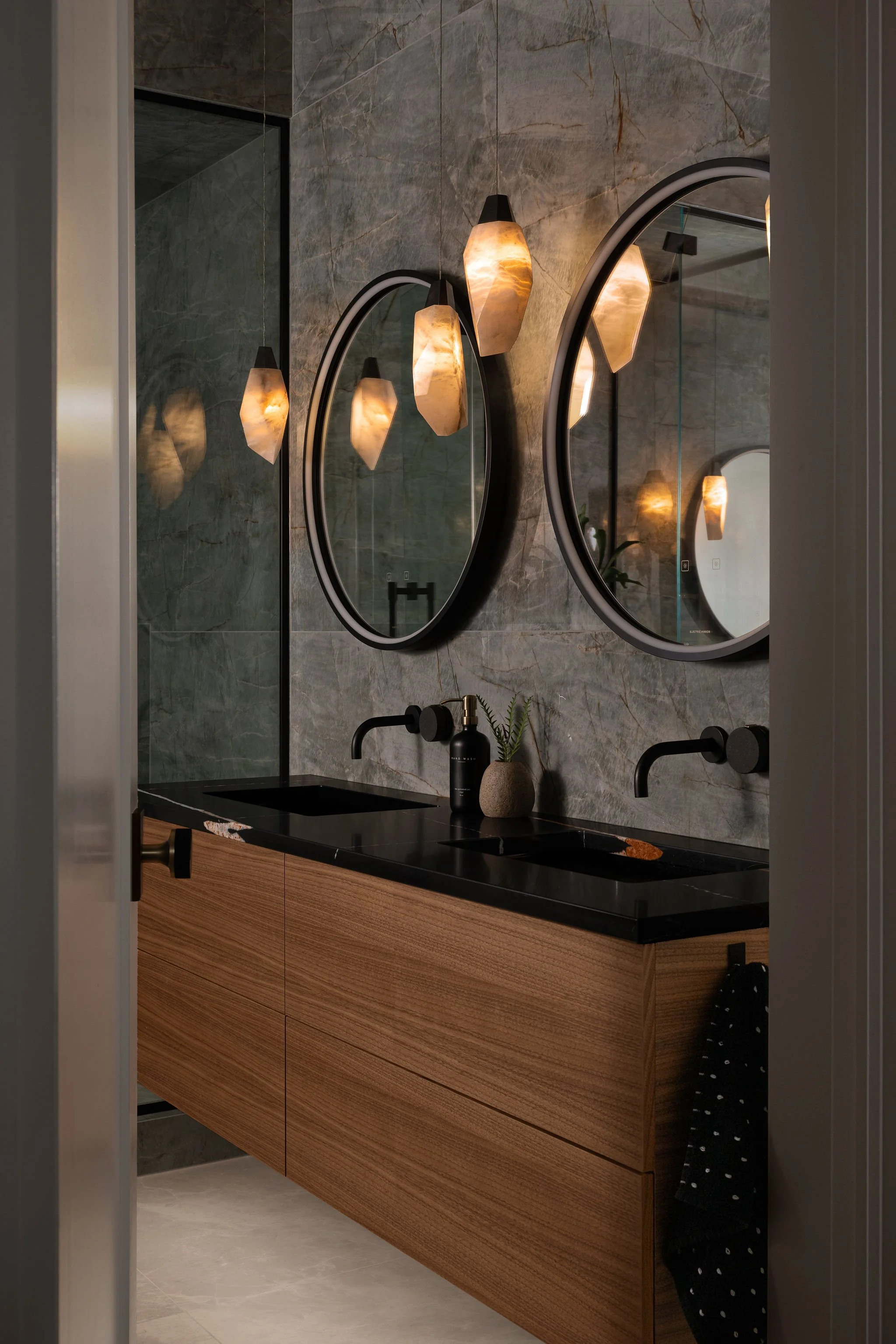 Primary ensuite bathroom with floating walnut double vanity, black countertop and sinks, round mirrors, black wall-mounted faucets, porcelain slab walls, and glowing alabaster pendant lights.