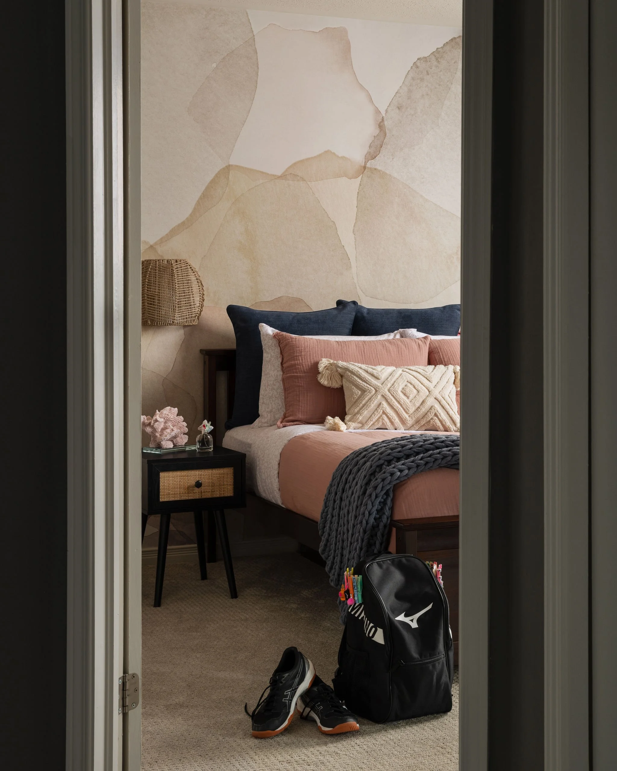 Teen bedroom viewed through an open doorway, featuring blush and coral bedding, a soft abstract mural wallcovering, woven wall sconce, cane nightstand, and school bag by the bed.