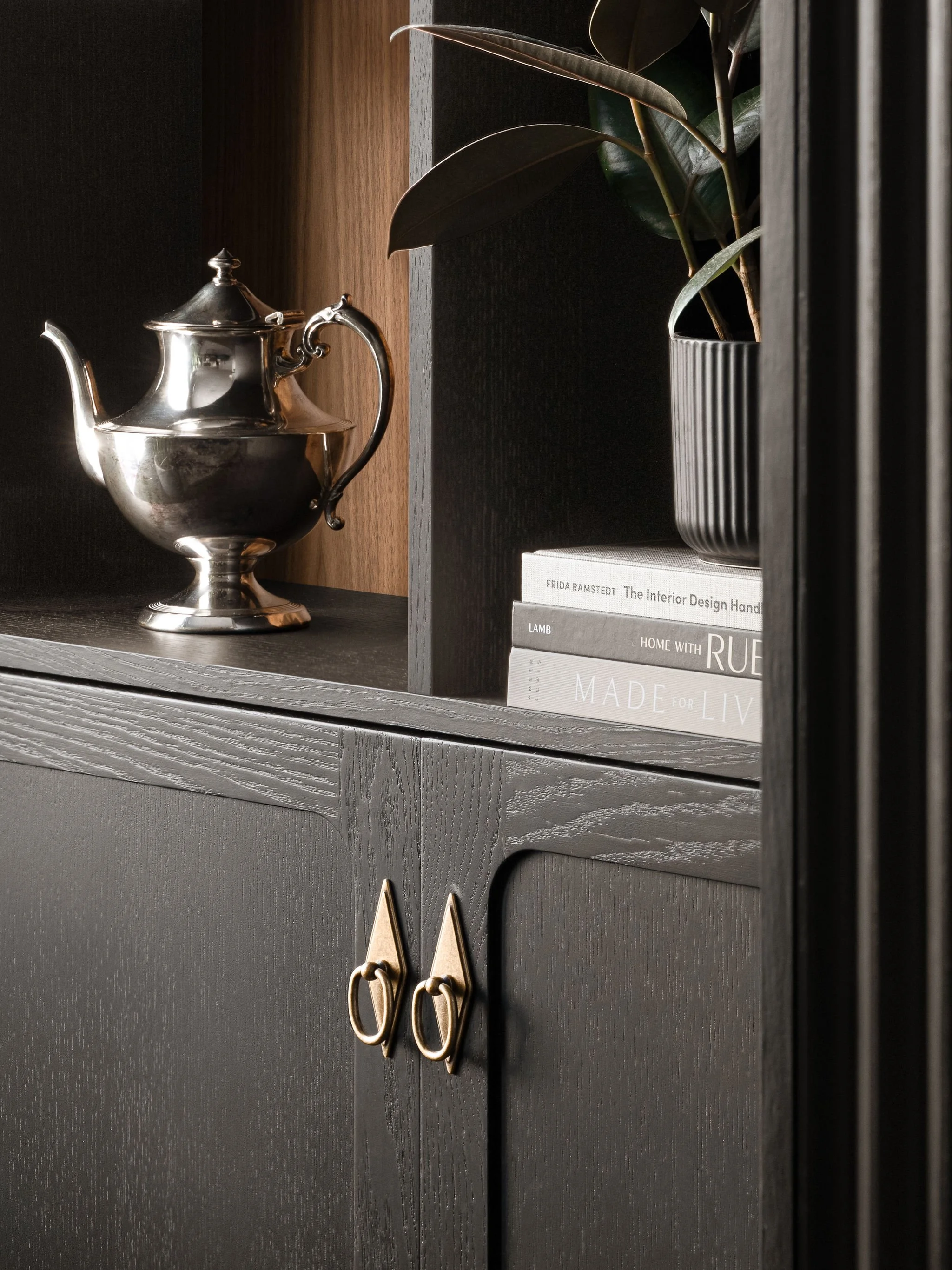 Close-up of custom black built-in cabinetry with brass hardware, silver teapot, stacked design books, plant, and warm wood shelving interior.