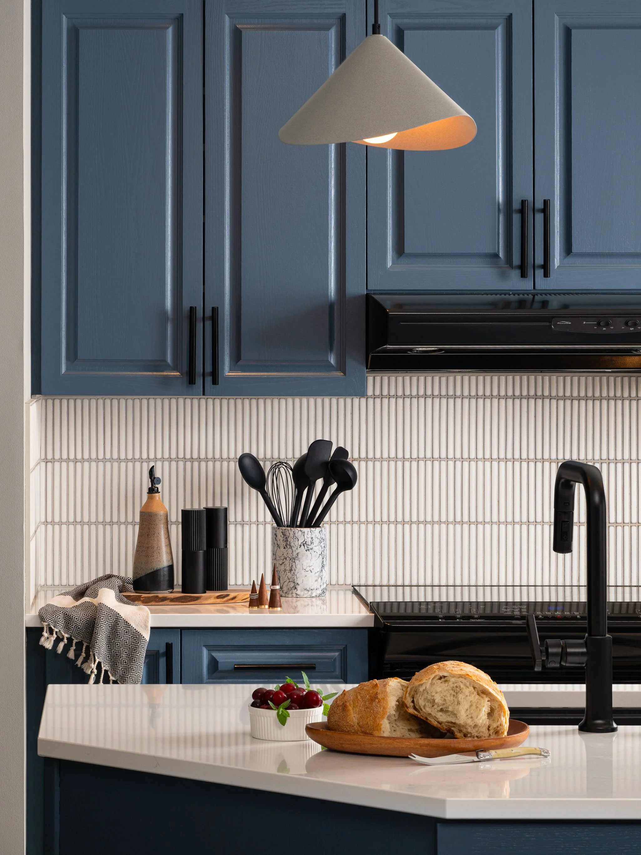 Blue kitchen cabinetry with black hardware, white vertical tile backsplash, white quartz countertop, black faucet, and sculptural cone pendant light.