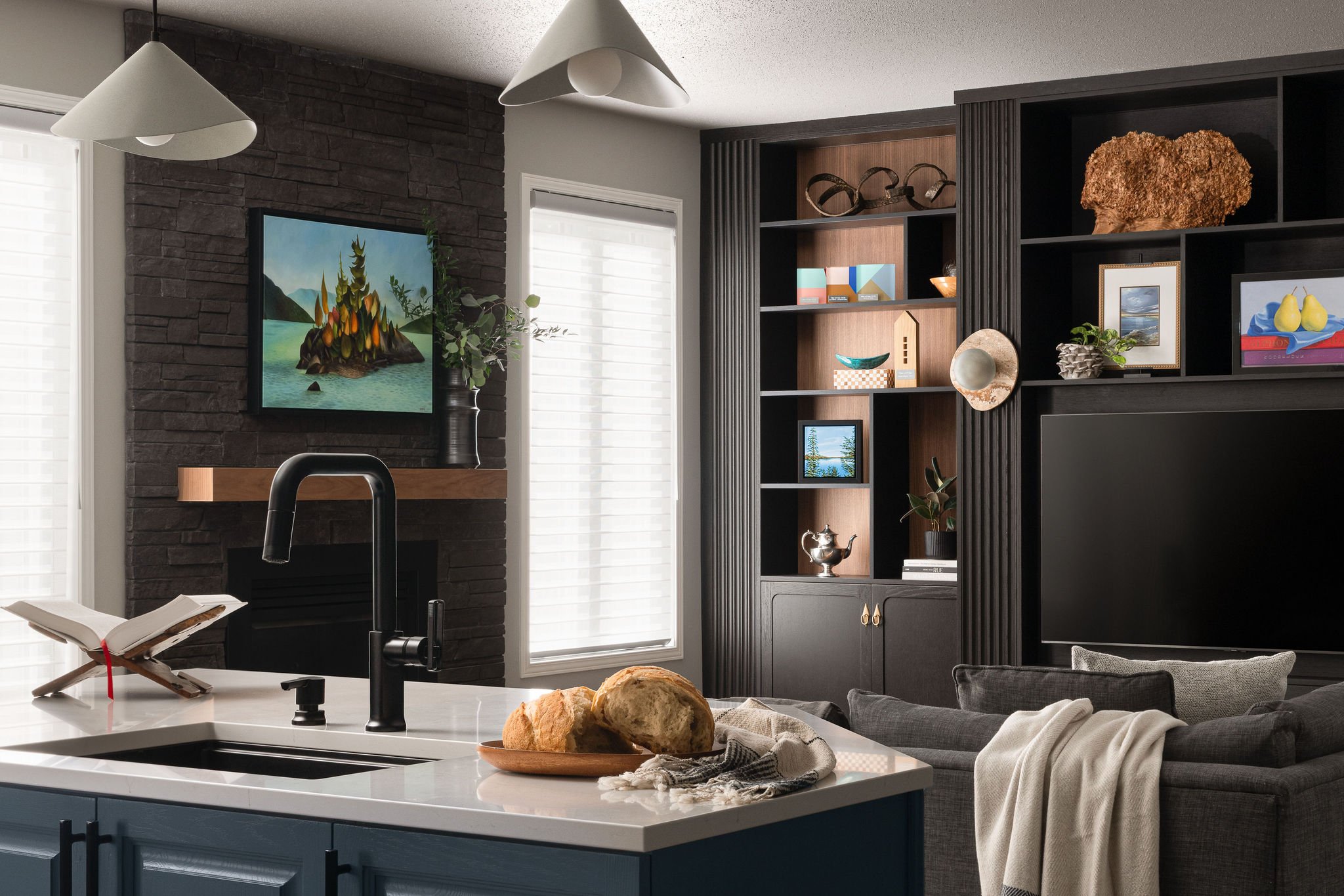 Open-concept kitchen and living room with blue cabinetry, white quartz island, black faucet, dark stone fireplace, and custom black built-in media wall with styled open shelving.