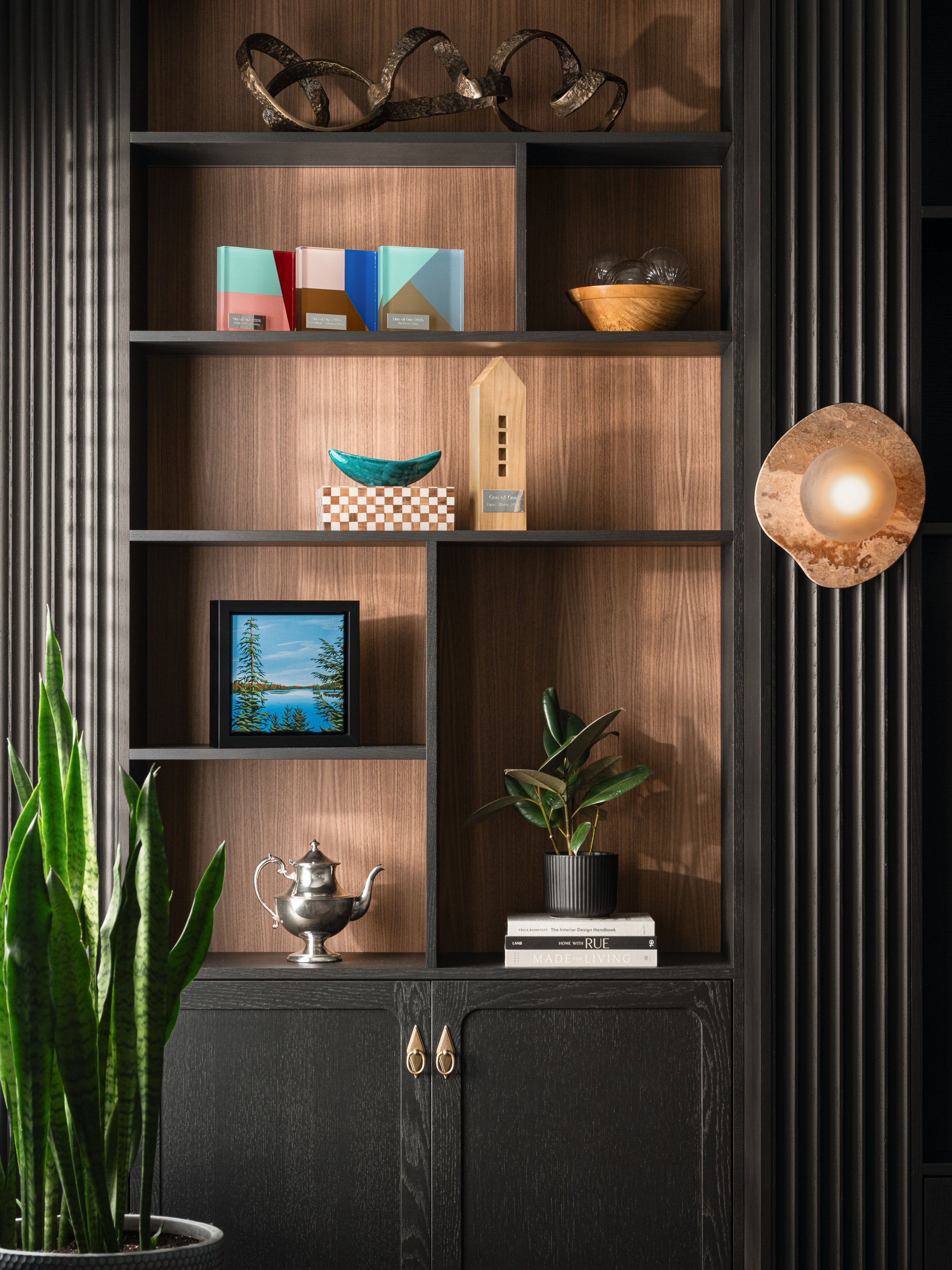 Styled open shelving in a custom black built-in with warm wood backing, integrated lighting, art, plants, decorative objects, books, and brass cabinet hardware.