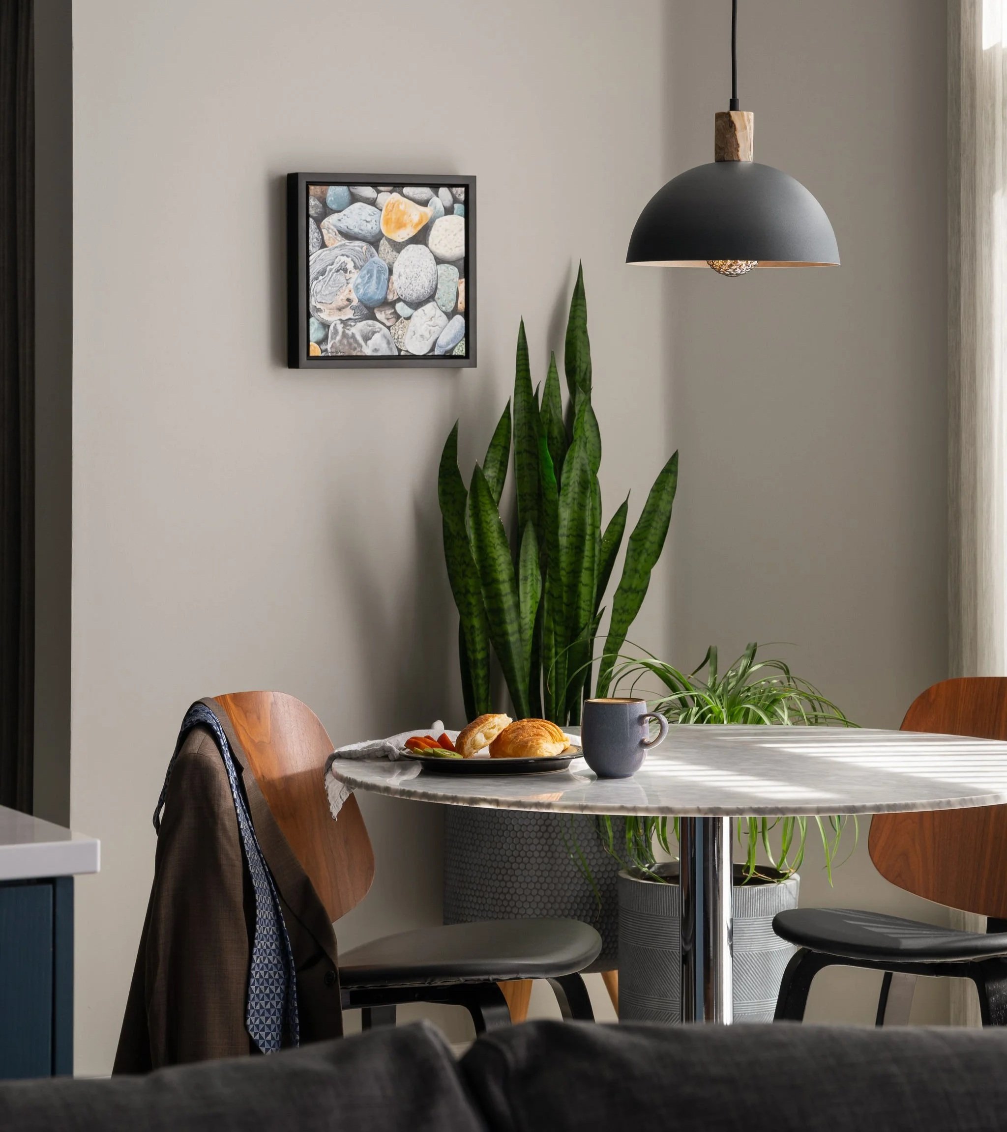 Small dining area with round marble table, wood and black chairs, black pendant light, layered plants, framed pebble artwork, and breakfast setting.