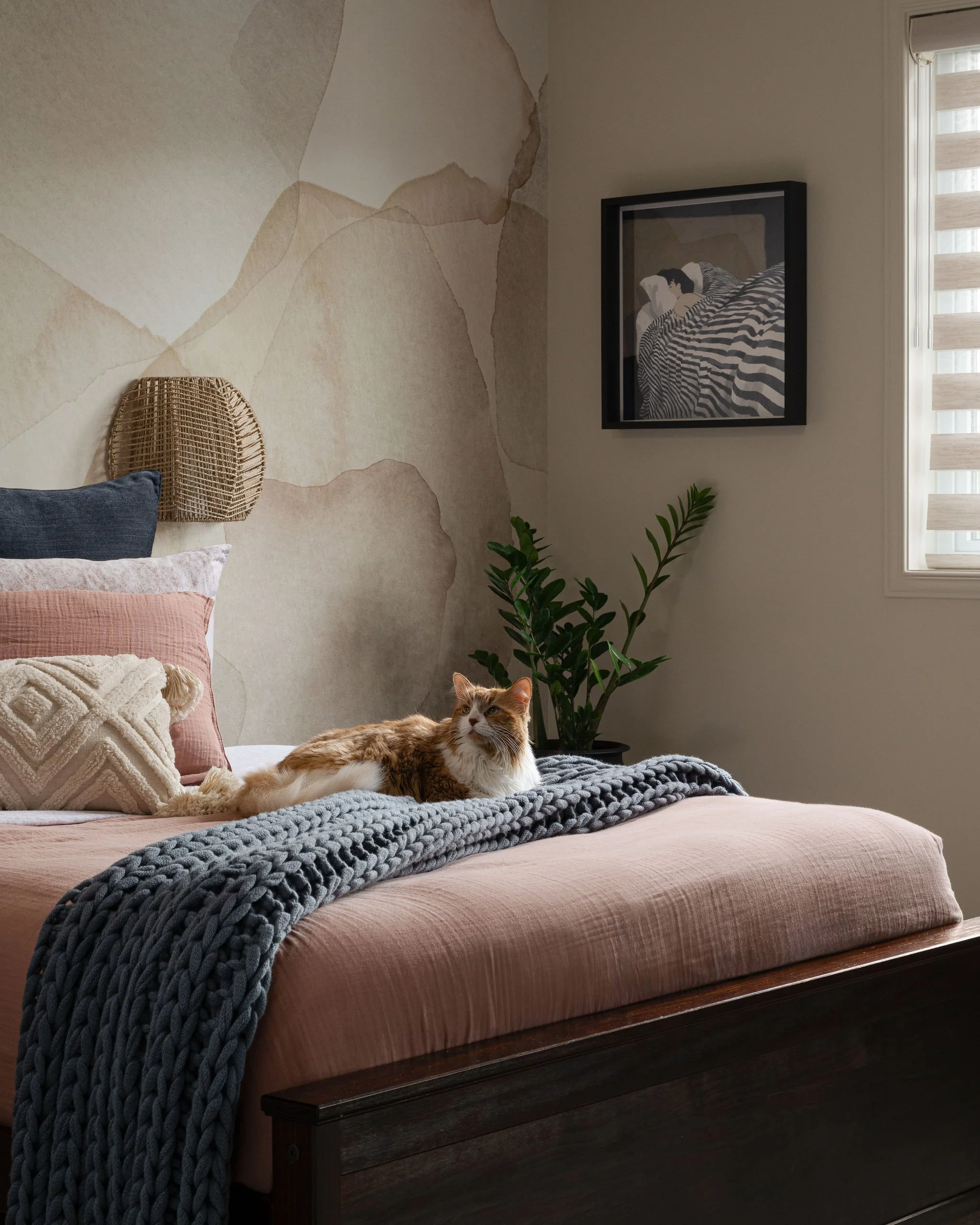 Layered teen bedroom with blush bedding, navy pillows, chunky charcoal throw, abstract mural wallcovering, woven wall sconce, framed art, plant, and cat resting on the bed.